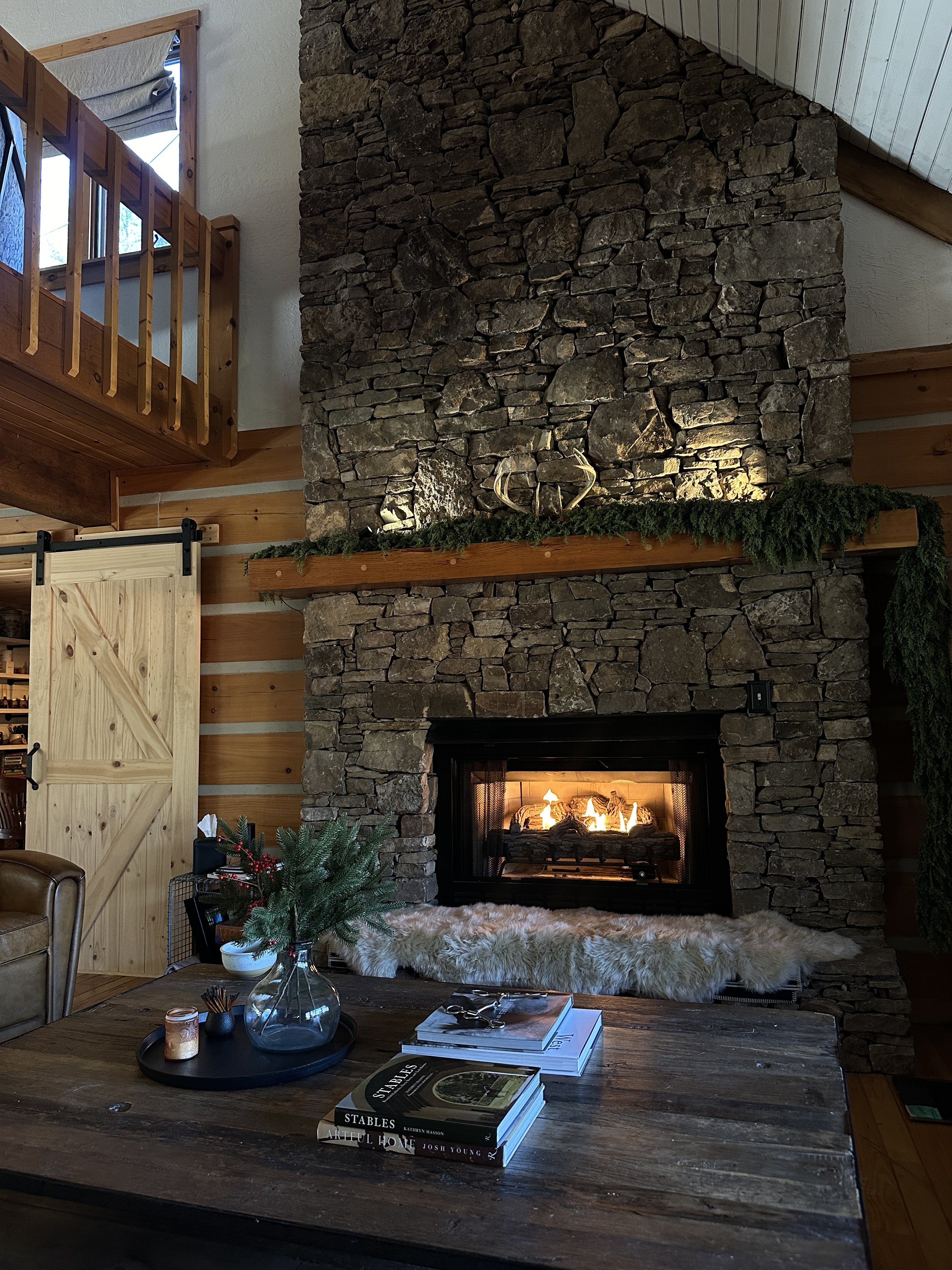 A cozy living room with a stone fireplace, a wooden mantel decorated with greenery and a metal deer antler sculpture, a fur throw at the hearth, a rustic wooden table with a small pine branch arrangement, books, and a magazine, and a wooden sliding d