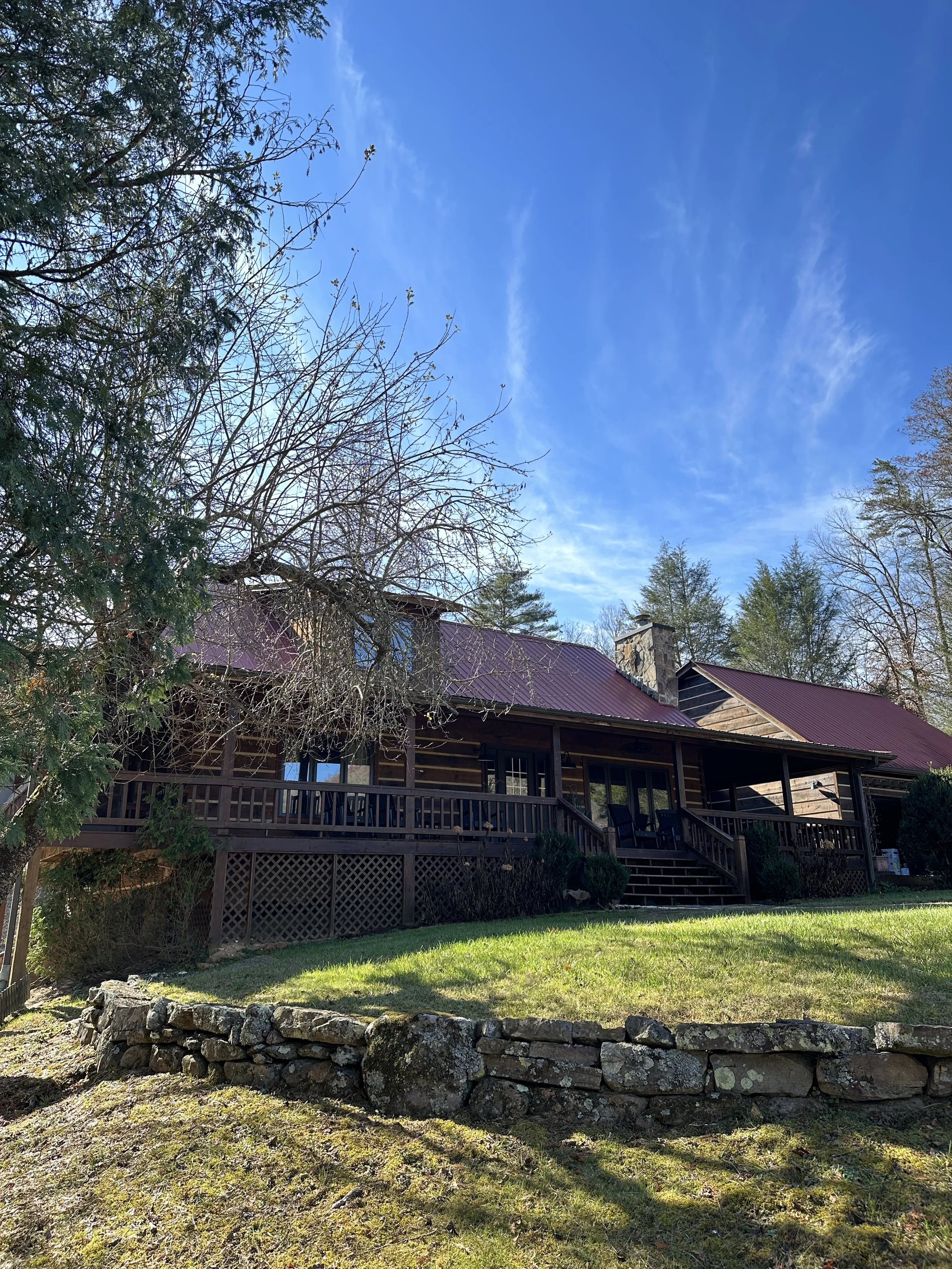 A rustic wooden house with a red metal roof, surrounded by trees and a grassy yard. The house has a porch with chairs, and a stone retaining wall is in the foreground under a blue sky.