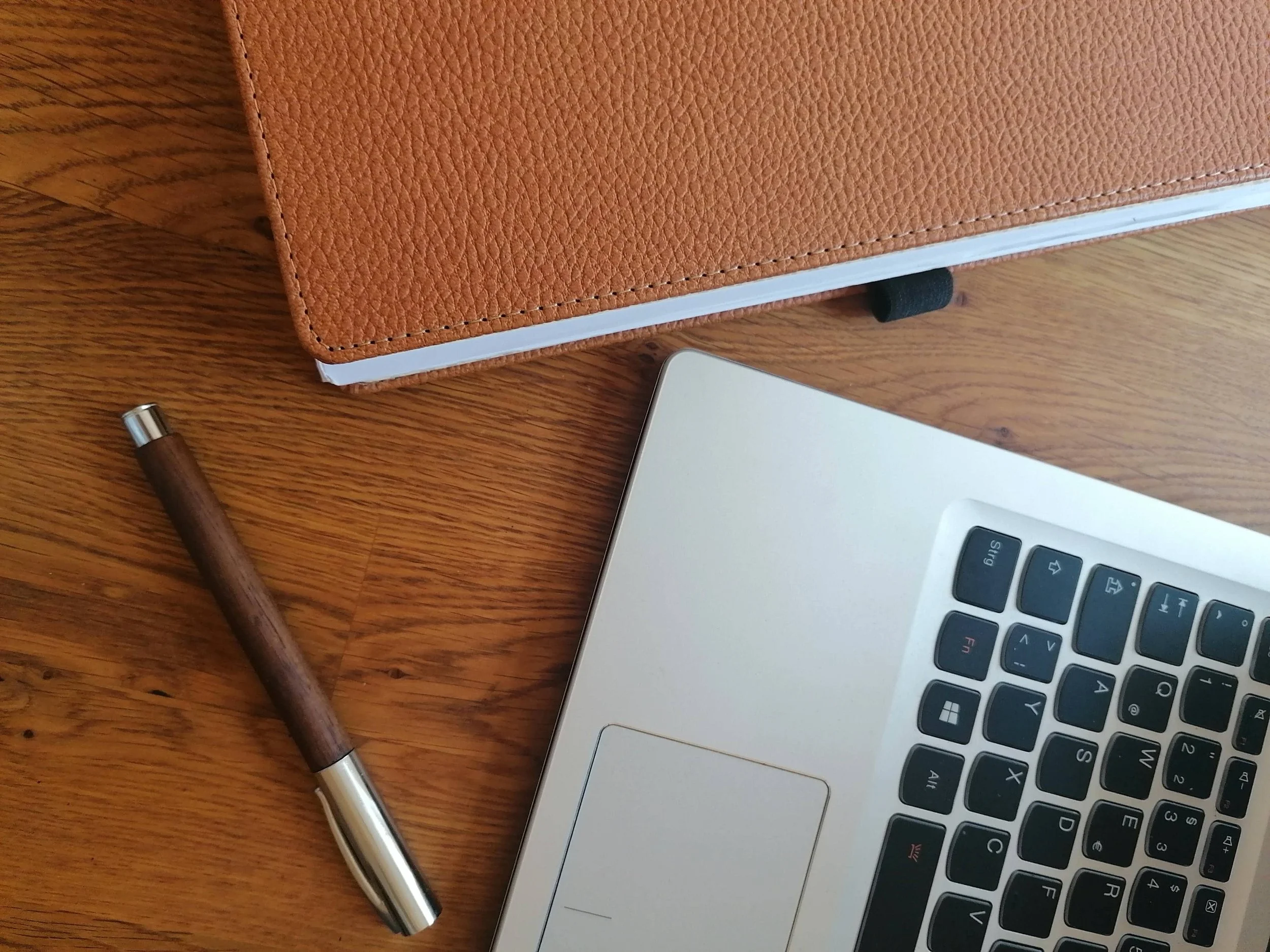 Ein braunes Notizbuch, ein Kugelschreiber, ein Laptop mit sichtbarer Tastatur auf einem Holzschreibtisch als Symbol für Fokus und Klarheit.