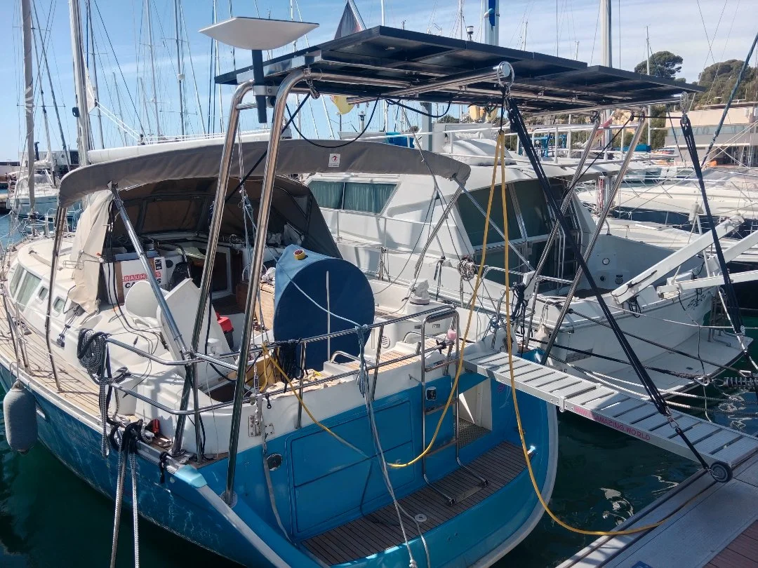 The image shows the stern and upper deck of a white sailboat with a solar panel mounted above. There is a ladder leading to the upper deck, a boat cover, and various equipment on the boat. The sky in the background is clear and blue.
