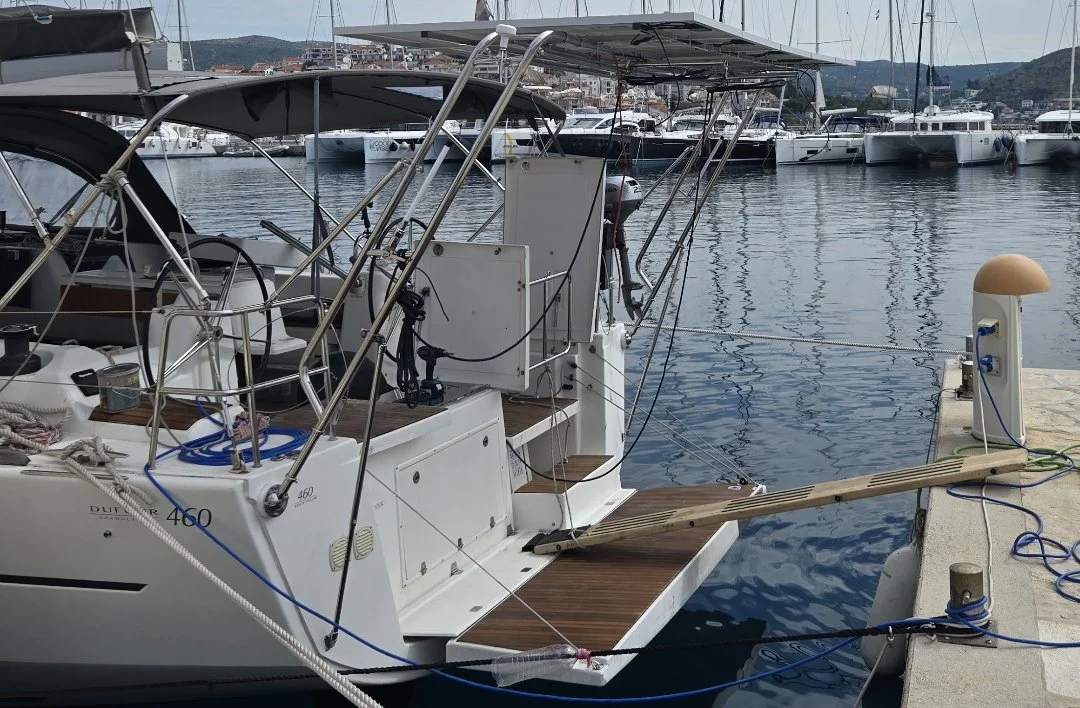 A white sailboat docked at a marina with other boats in the background, featuring a wooden swim platform and covered cockpit area.