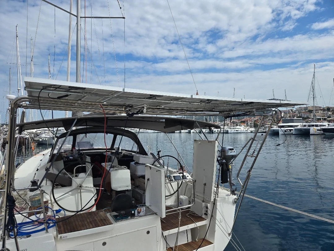 A sailboat docked in a marina with a solar panel on top, multiple steering wheels, and various equipment on the deck, with other boats and a hill in the background under a partly cloudy sky.