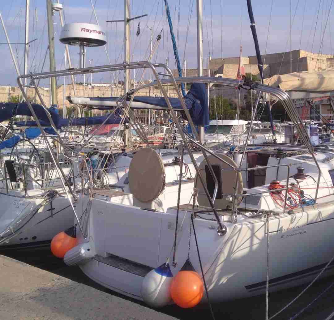 A docked sailboat with a white hull, orange and black fenders hanging from the sides, and a metal railing. The sailboat is equipped with antennas and equipment, with other boats and buildings in the background.
