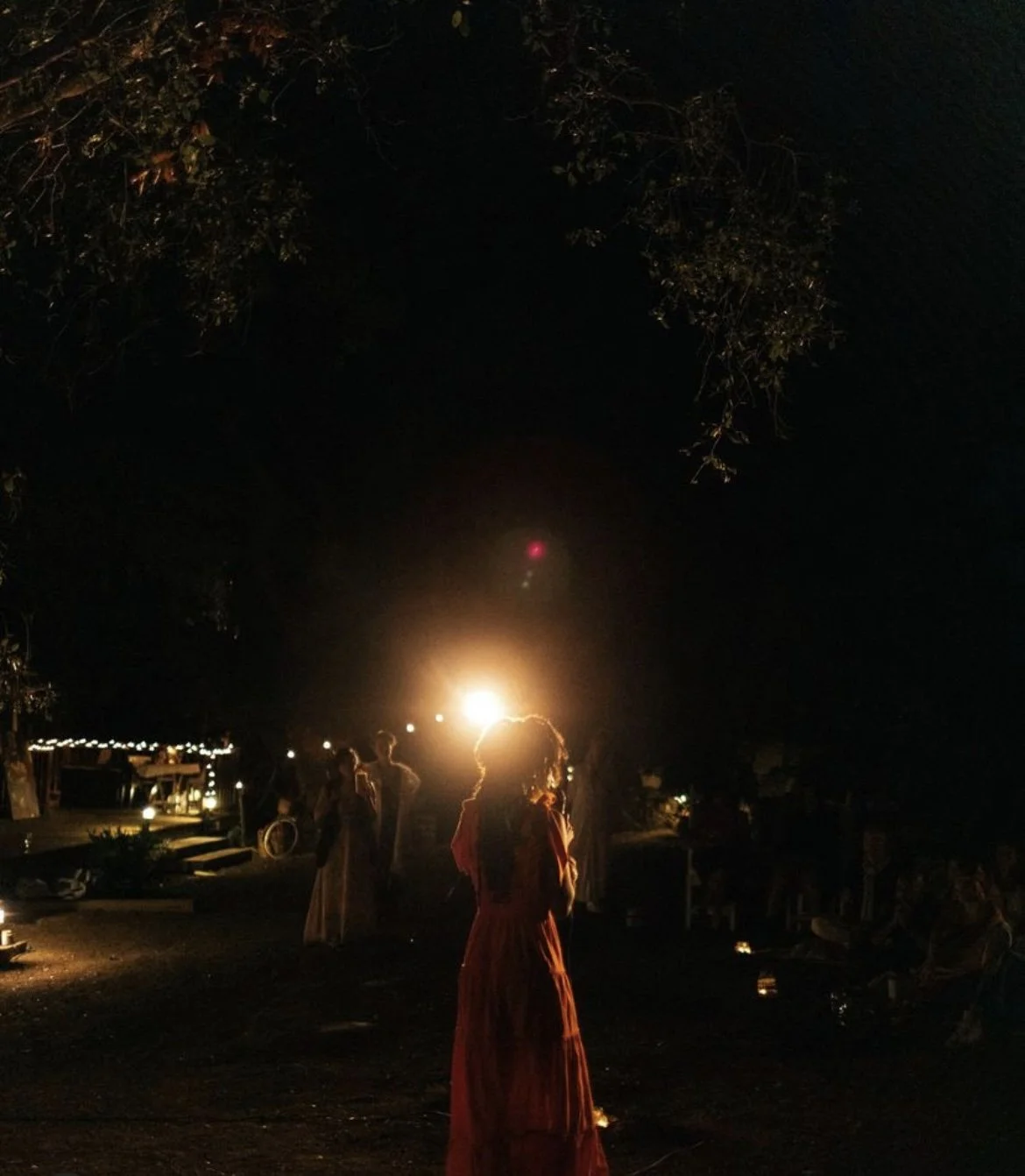People gathered outdoors at night with a bright light source behind them, some sitting and some standing. The scene is dark, with silhouettes of trees and a pathway.