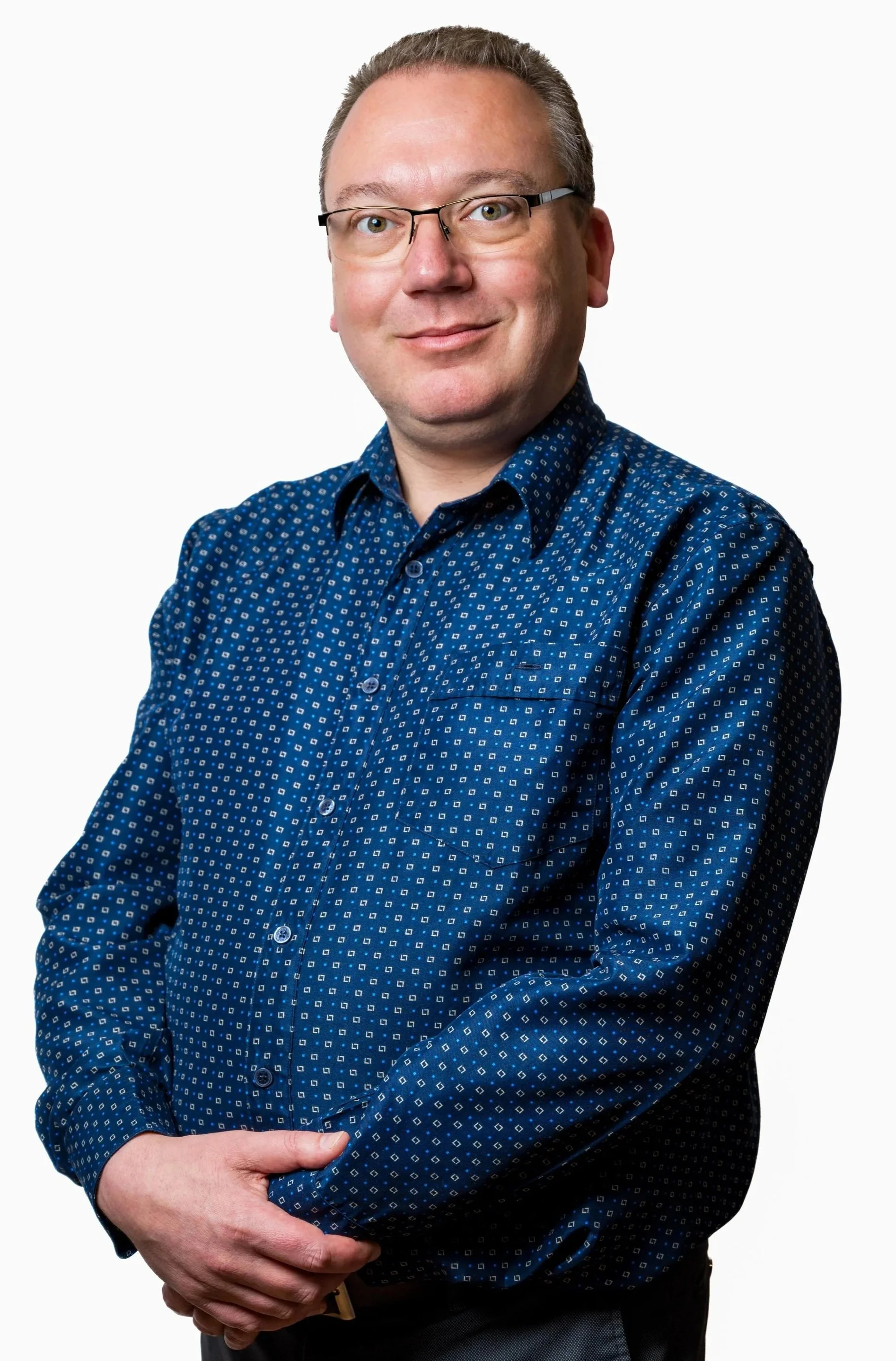 A man in glasses smiling, wearing a dark blue patterned shirt, with his arms crossed and hands resting on his waist.