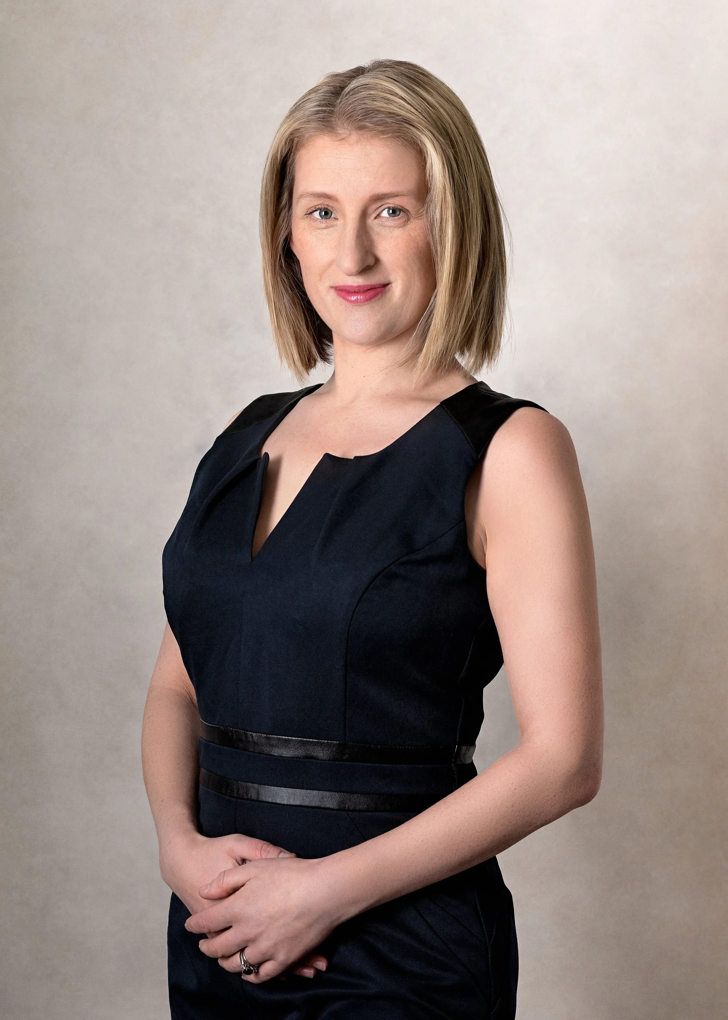 A woman with shoulder-length blonde hair, wearing a sleeveless black dress, looking at the camera with a slight smile, standing against a plain, light-colored background.