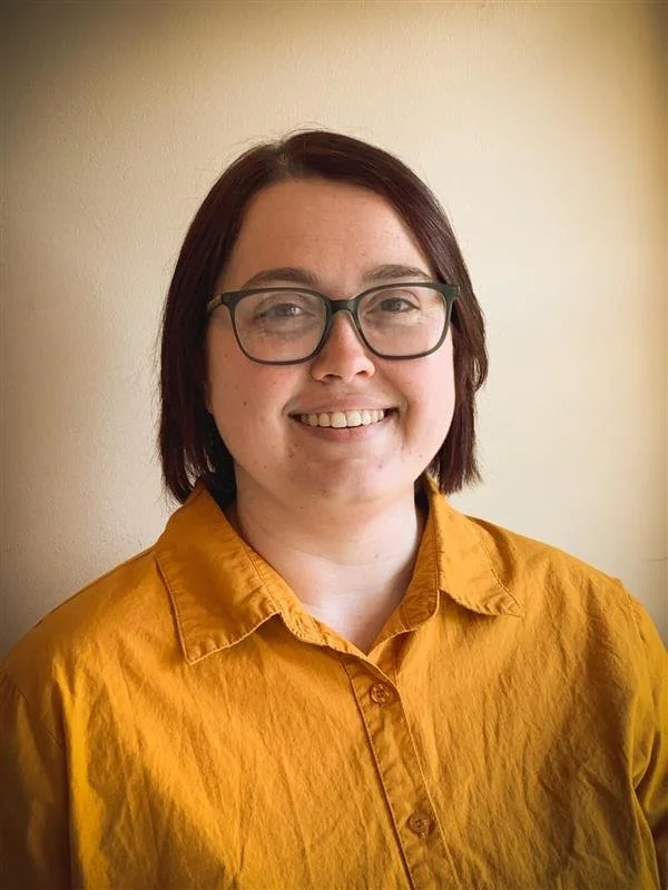 A woman with short brown hair and glasses smiling at the camera, wearing a mustard yellow shirt, standing against a beige background.