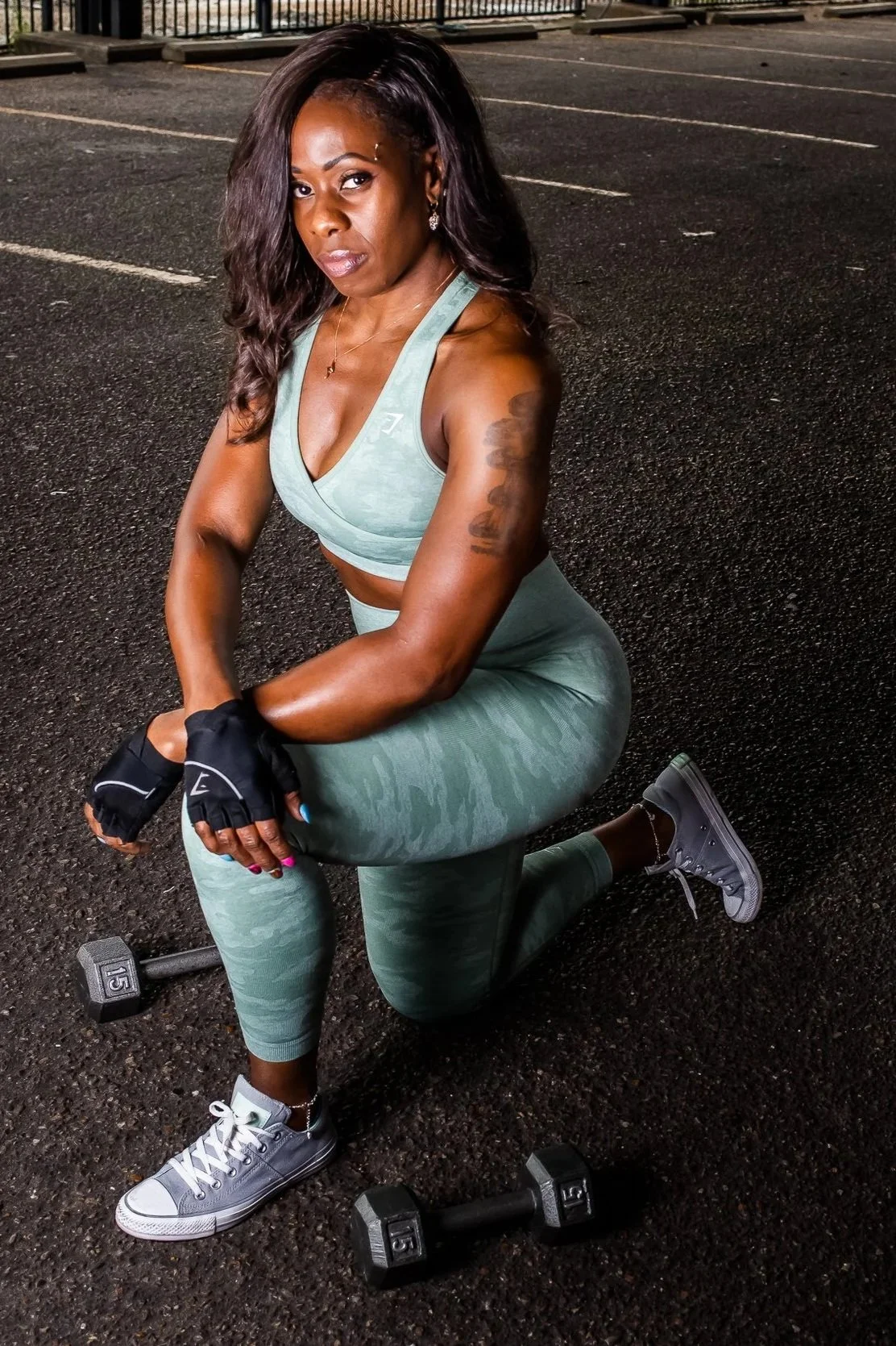 A woman wearing workout clothes, kneeling on a gym floor, holding dumbbells, with a determined expression.