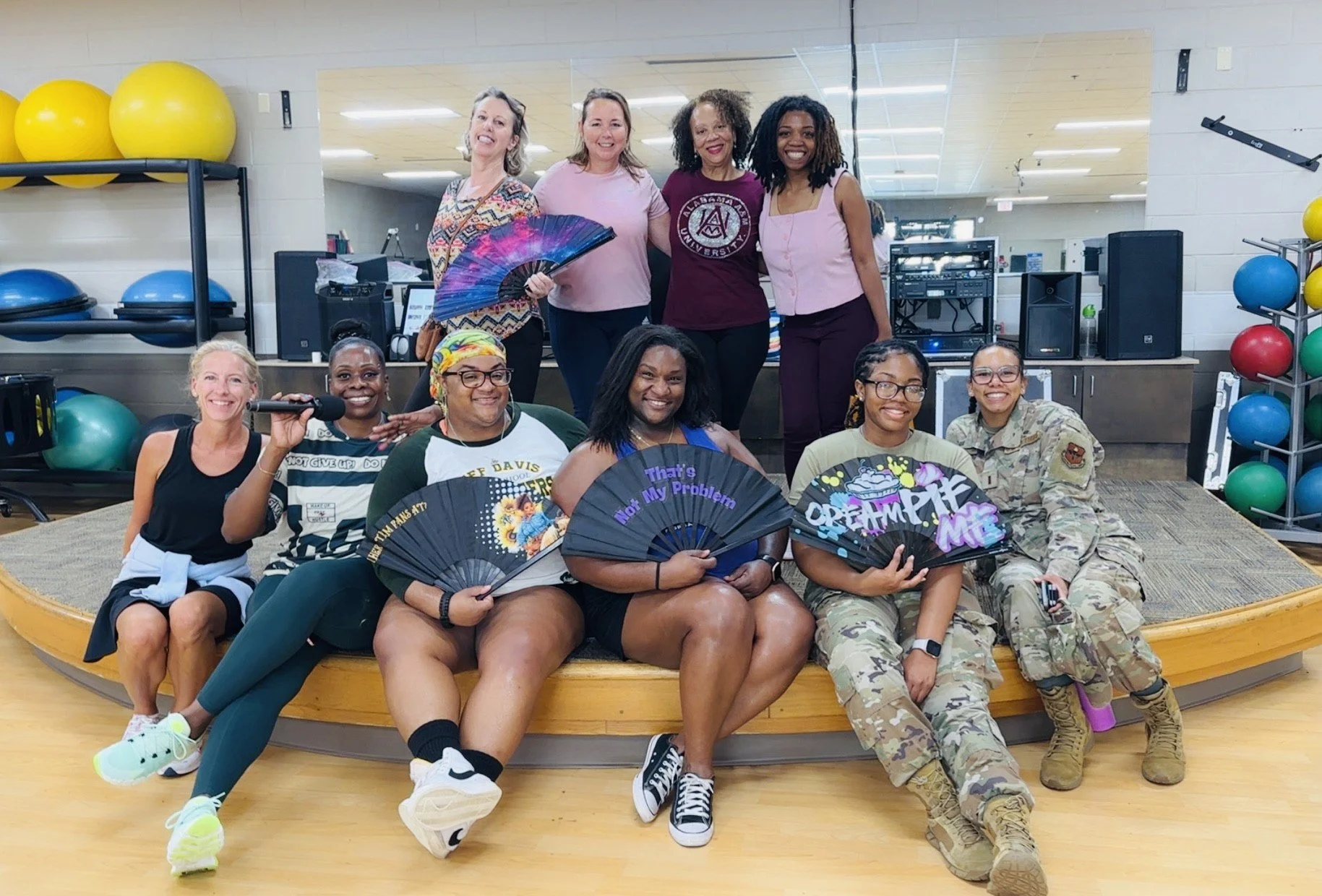A group of women, some in casual wear, others in military uniform, sitting and standing on a wooden stage in a gym or fitness room. They are holding decorated fans with messages like 'That's My Problem' and 'Dream Big.' The back row has women smiling, with one holding a colorful fan. The gym background features exercise balls, mirrors, and sound equipment.