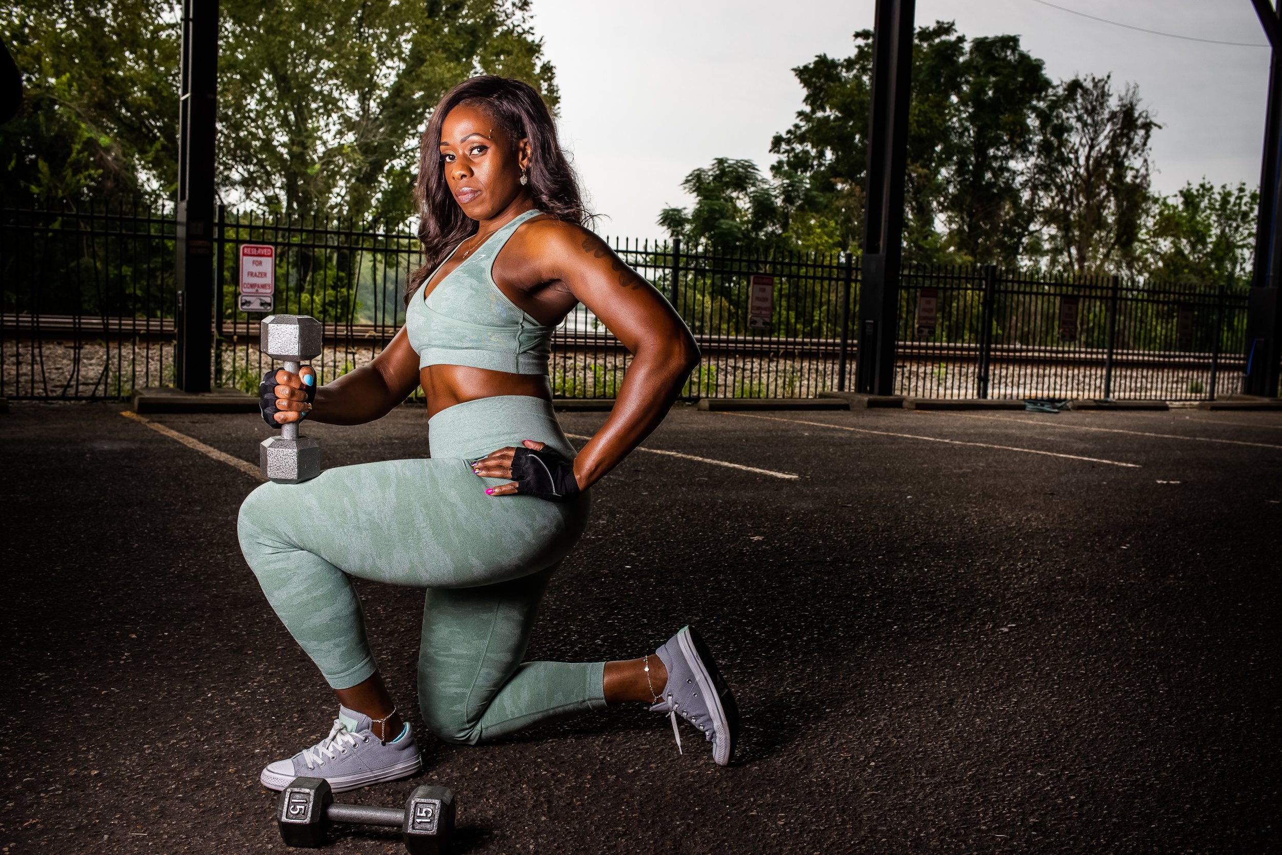 A woman kneeling on one knee in a parking lot, holding a dumbbell in her right hand, wearing grey workout clothes and sneakers, with trees and a fence in the background.