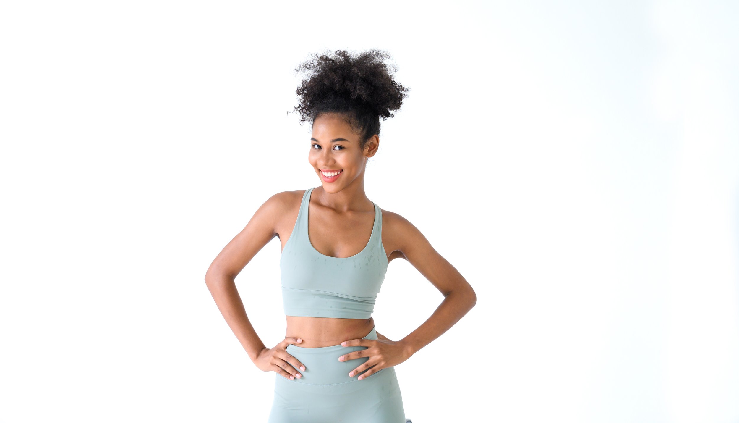 A young woman in athletic wear smiling with hands on hips against a white background.