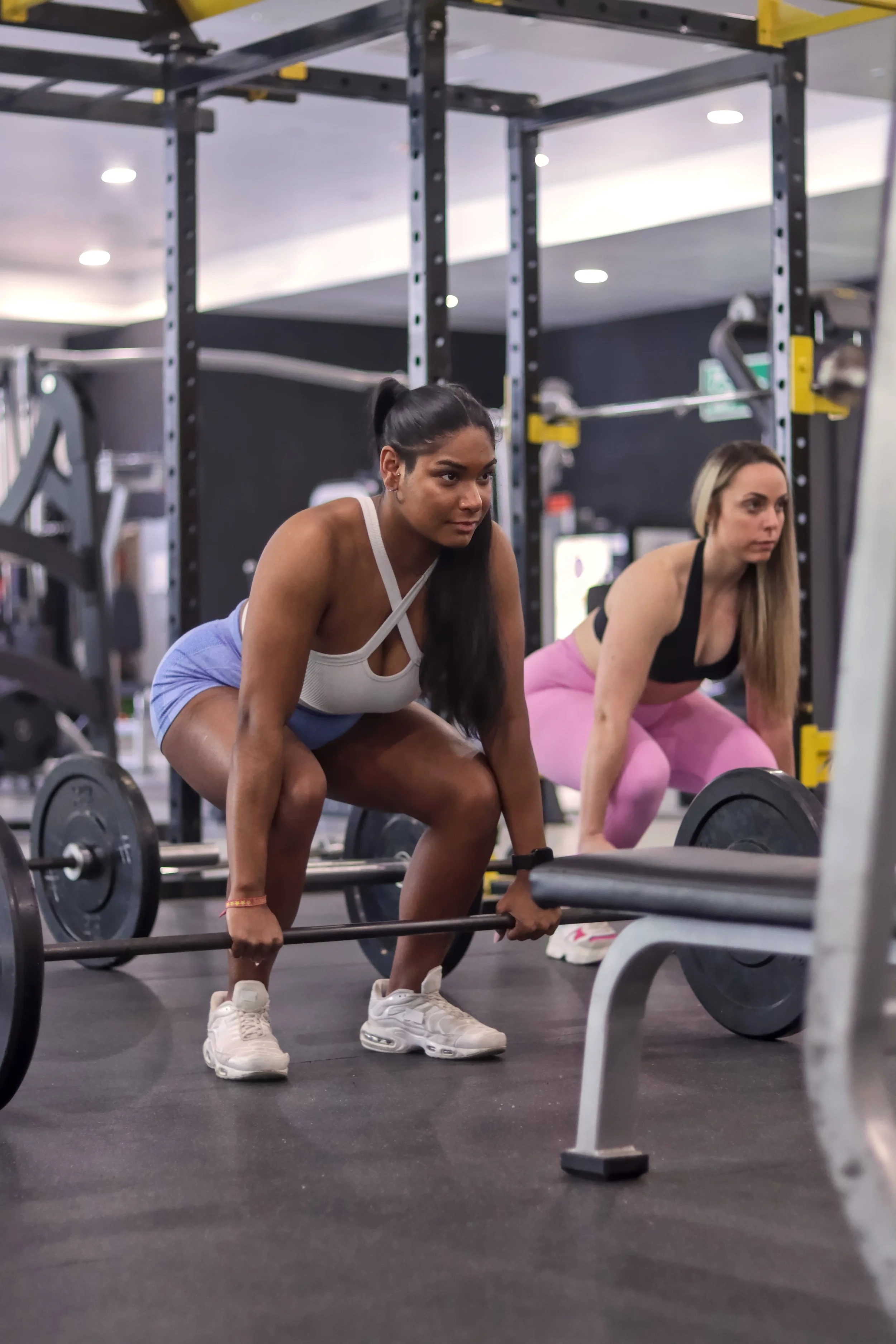 Two women lifting barbells in a gym, bent over with focused expressions, black workout racks in background.