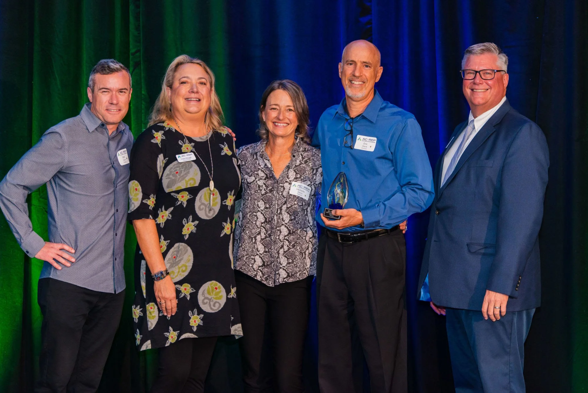 Group of six professionally dressed people standing together on stage, with one man holding an award, against a backdrop of multicolored curtains, at an awards event.