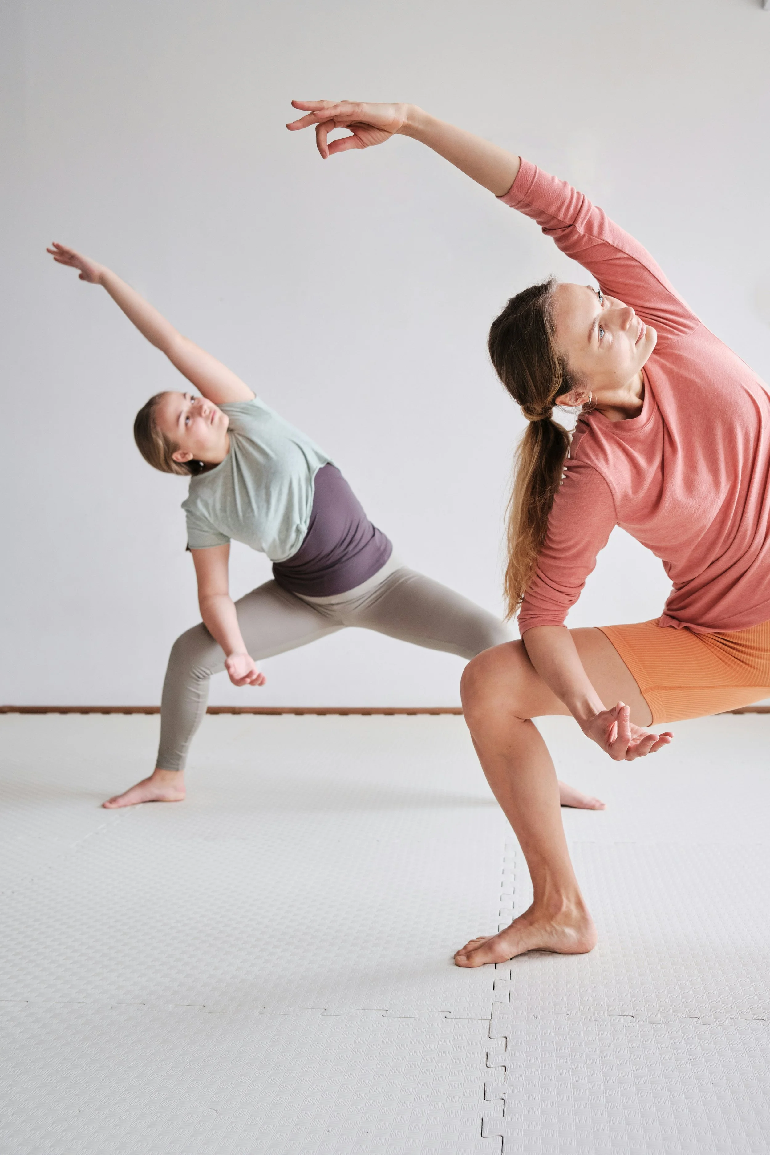 Two women practicing yoga in a studio, both in a side lunge position with arms extended overhead, one on the left and one on the right, on a white textured mat.
