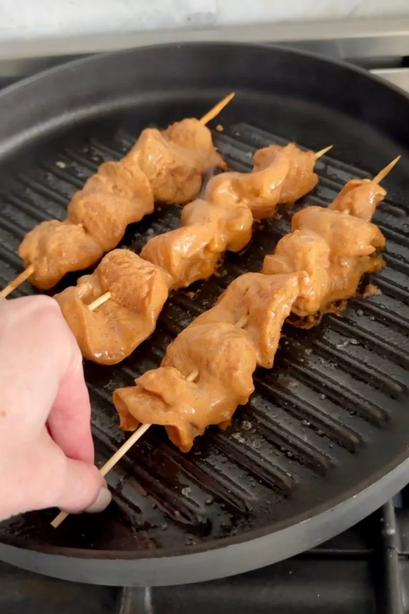 Close-up of grilled chicken satay skewers garnished with crushed peanuts and served on a plate with lime wedges and sliced red chili peppers.