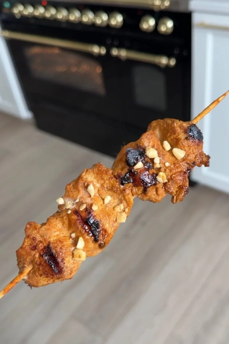 A hand holding a chicken satay skewer topped with crushed peanuts.
