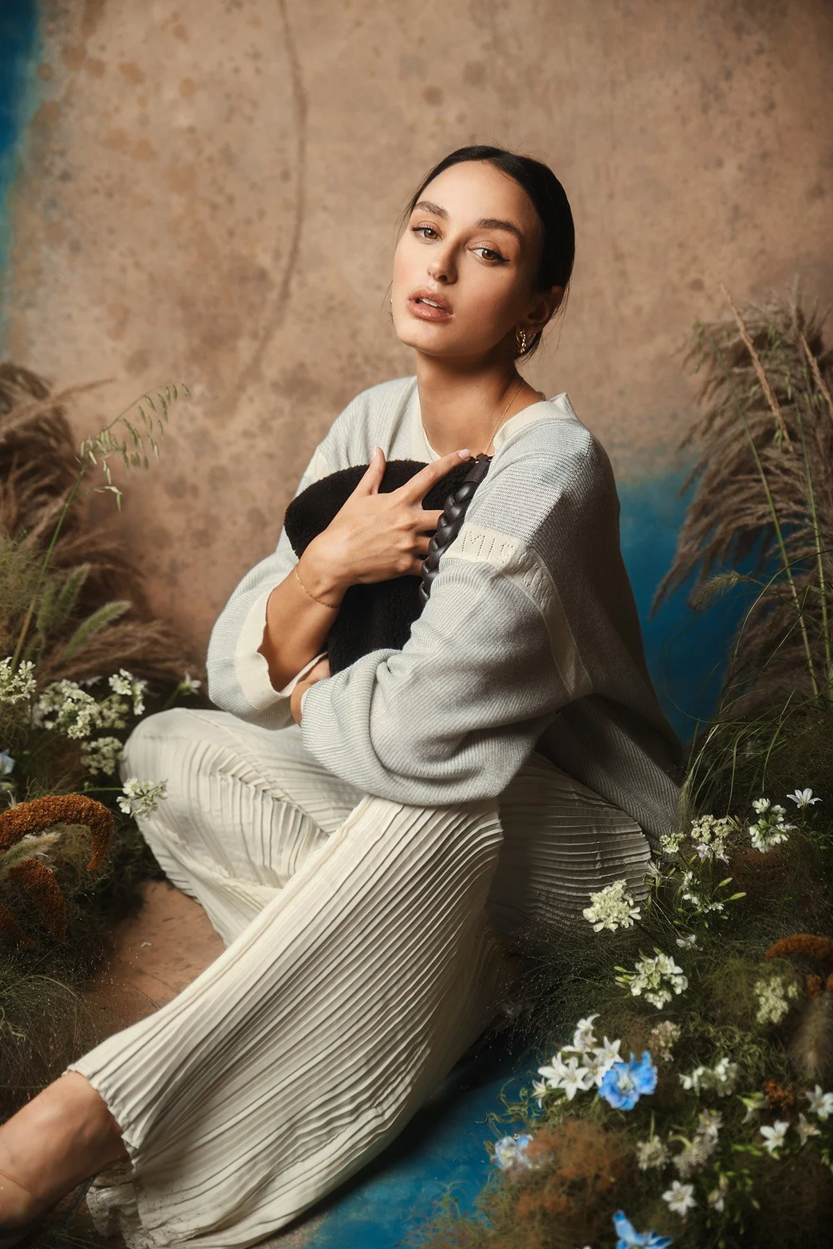 A woman with dark hair and fair skin sitting in a natural setting with flowers and grasses, wearing a light-colored sweater and pleated pants, looking at the camera with her hand on her chest.