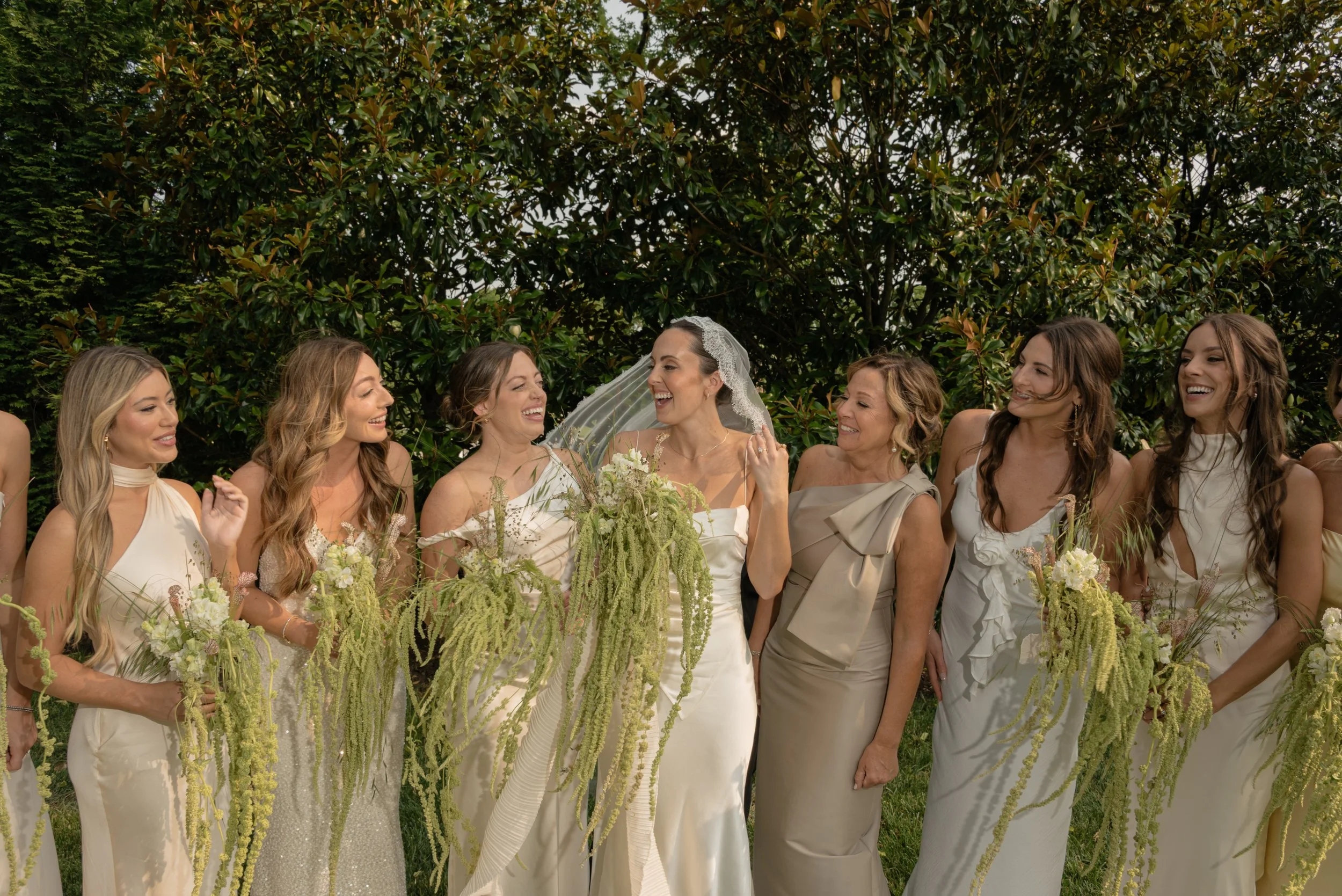 A group of women at a wedding, standing outdoors in front of trees, wearing elegant dresses, and holding flowers, smiling and laughing.