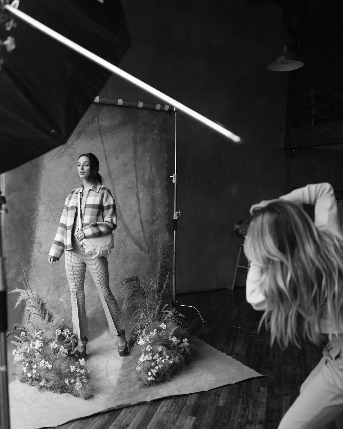 Black and white photo of a woman in stylish checkered pants and a casual jacket posing for a photoshoot in a studio with floral arrangements at her feet.