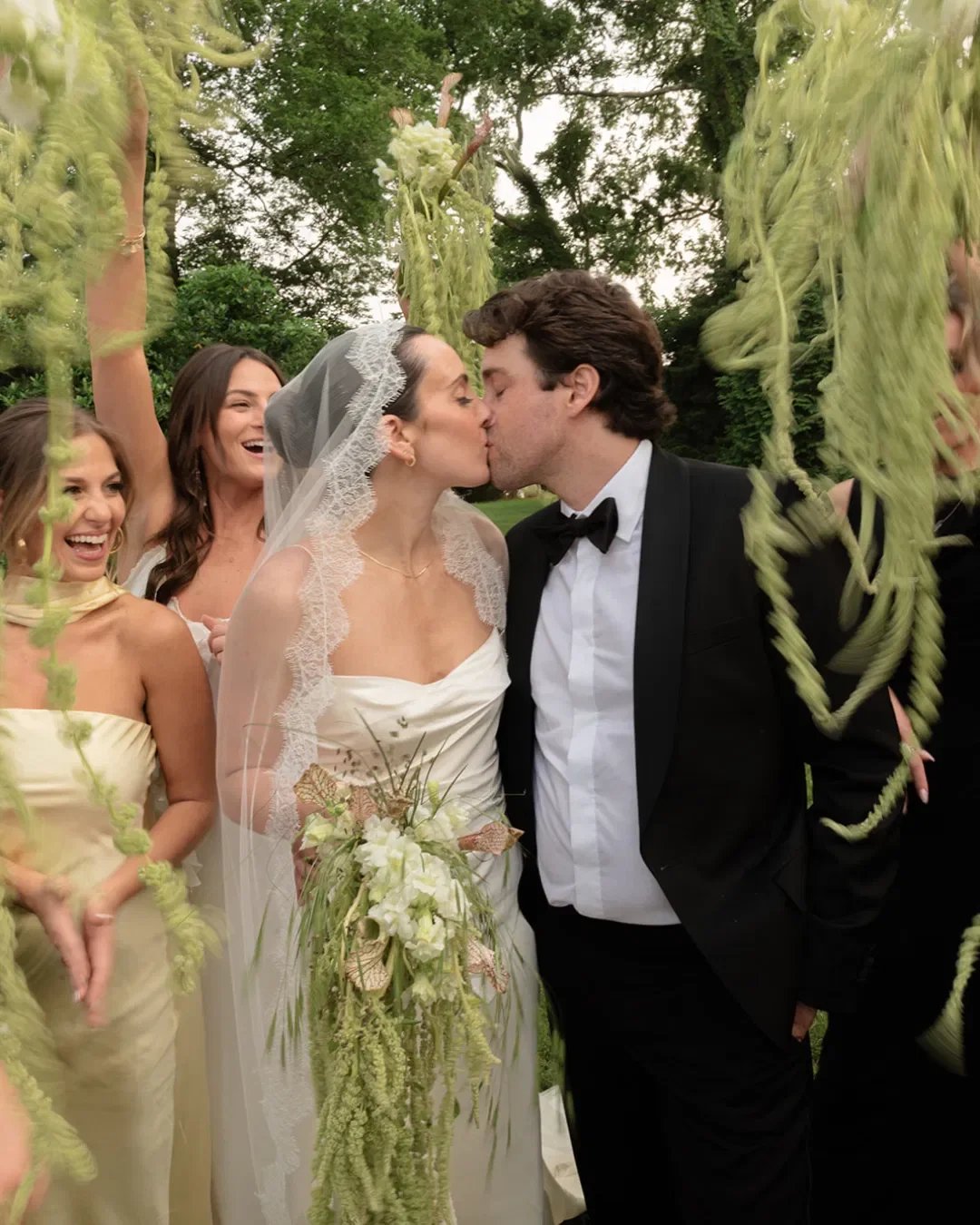 A wedding couple is kissing in the center while surrounded by friends outdoors, with greenery and hanging vines in the background.