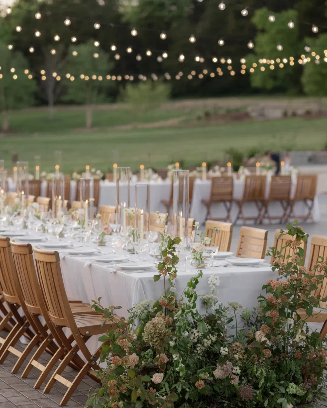 Outdoor wedding or event setup with long rectangular table covered with white tablecloth, decorated with candles, flowers, and glassware. String lights hang overhead, and wooden chairs surround the table. Green trees and grass in the background.