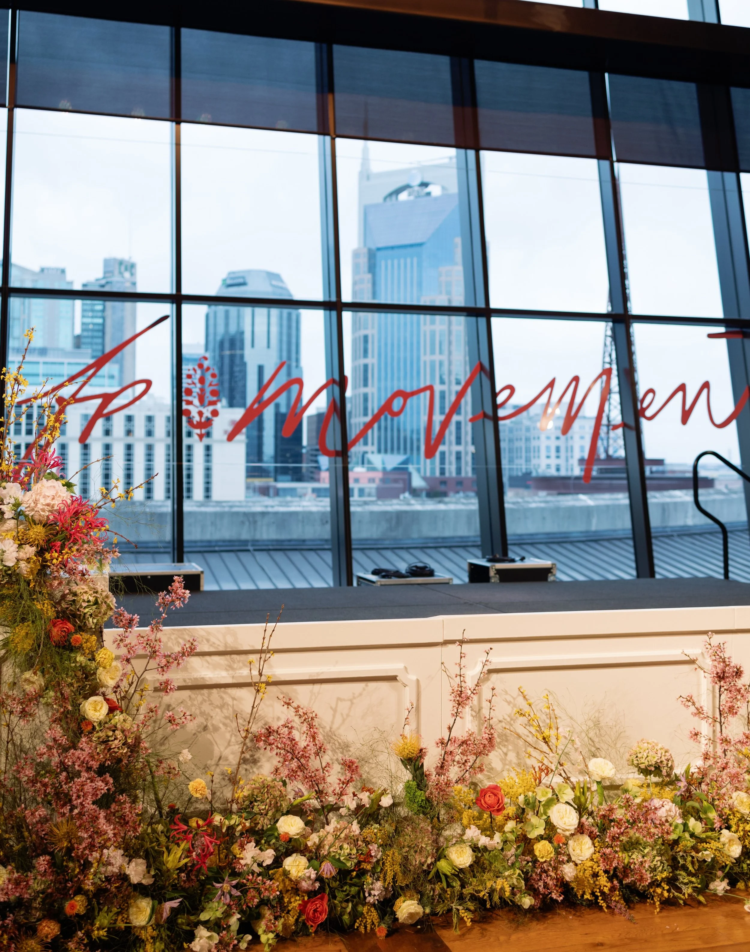 A large indoor floral display with a backdrop of tall city buildings, including a prominent skyscraper with a pointed top, visible through large windows. The flowers are colorful, featuring pink, white, yellow, and orange blooms, arranged along a whi