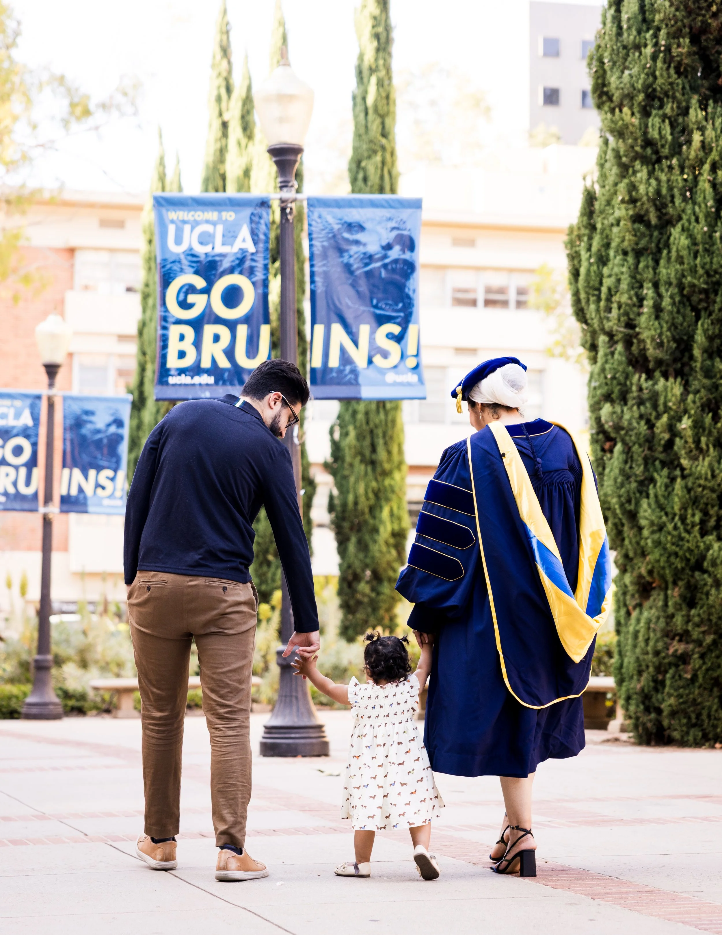 Two adults, a girl, and a baby walking together in a park. The man is wearing glasses, a navy sweater, tan pants, and tan shoes. The woman is in a graduation gown with a blue robe and cap, and black high-heeled sandals. The girl, holding hands with t