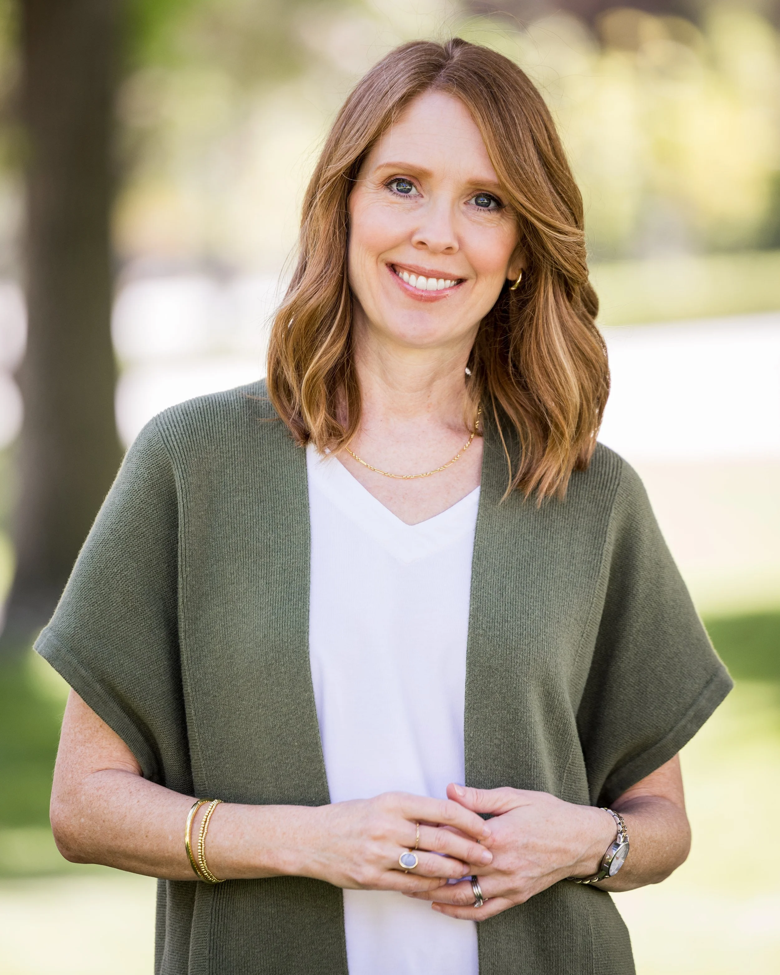 A woman with reddish-brown hair wearing a green blazer, white top, gold jewelry, and a watch, smiling outdoors with blurred trees in the background.