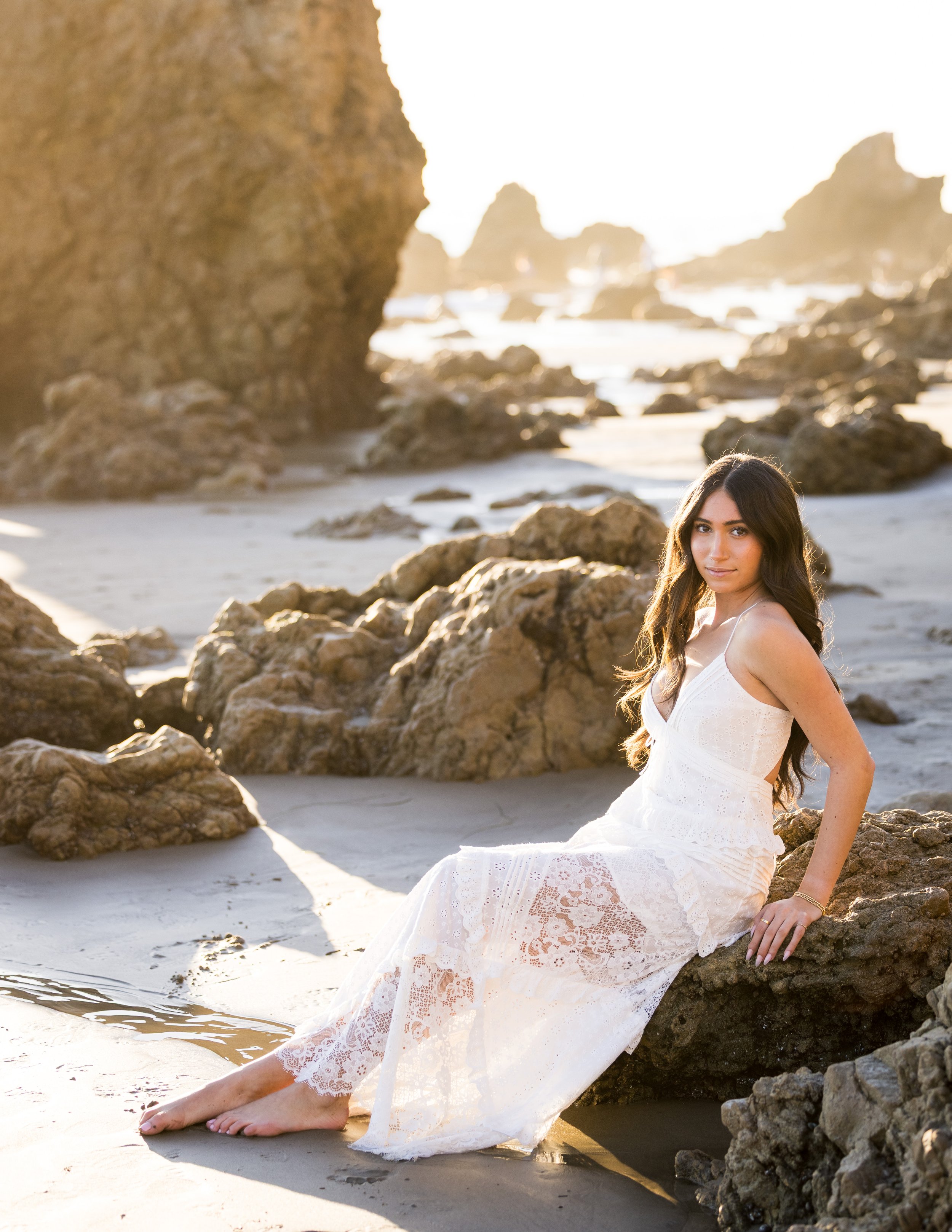 A young woman in a white lace dress sitting on rocks at the beach, with her feet in the sand and water, during sunset or sunrise.