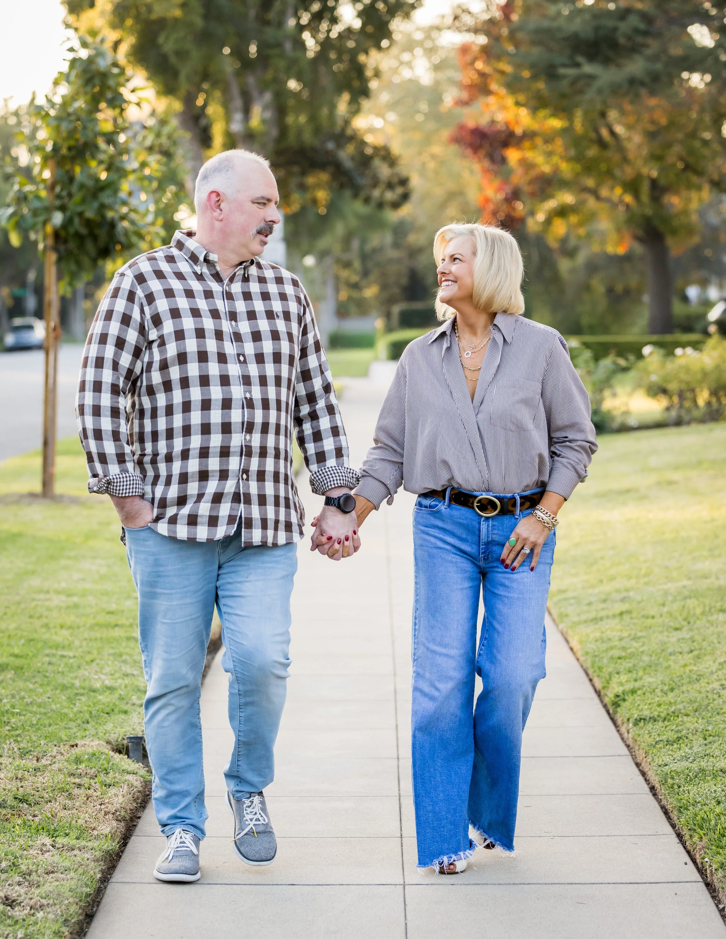 A couple walking hand in hand on a sidewalk in a park during sunset, smiling and looking at each other, surrounded by trees with fall foliage.