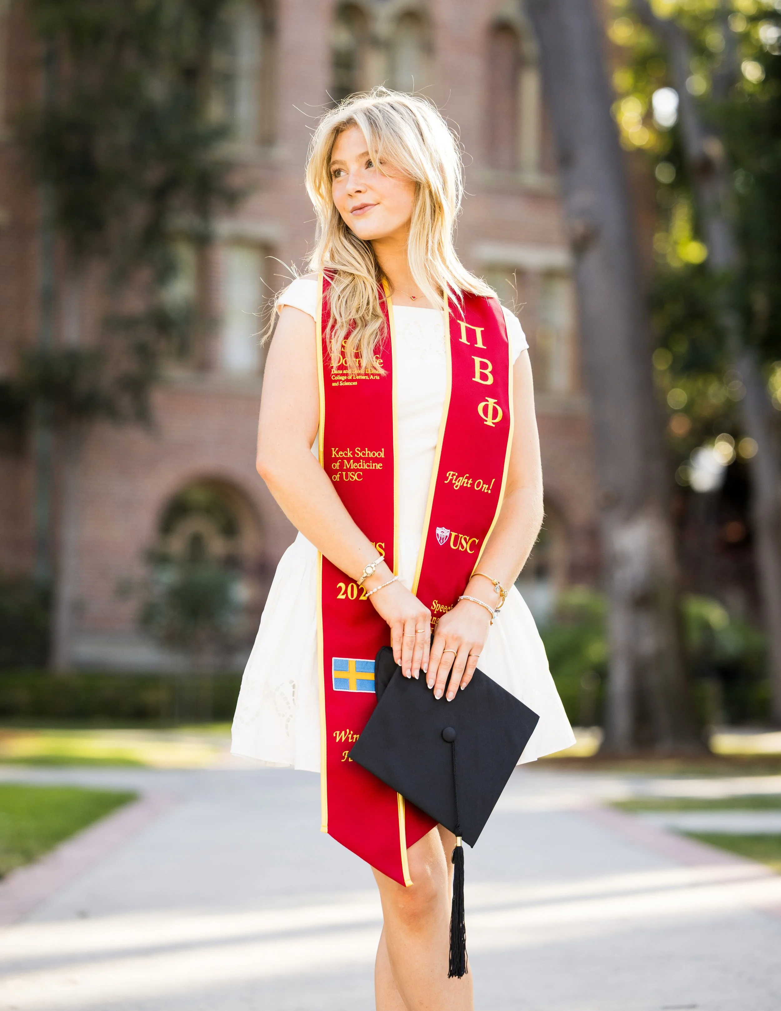 A young woman in a white dress wearing a red graduation stole holding a black cap and gown. She is outdoors, standing on a sidewalk with trees and a brick building in the background. The stole has Greek letters, the USC logo, and congratulatory messa