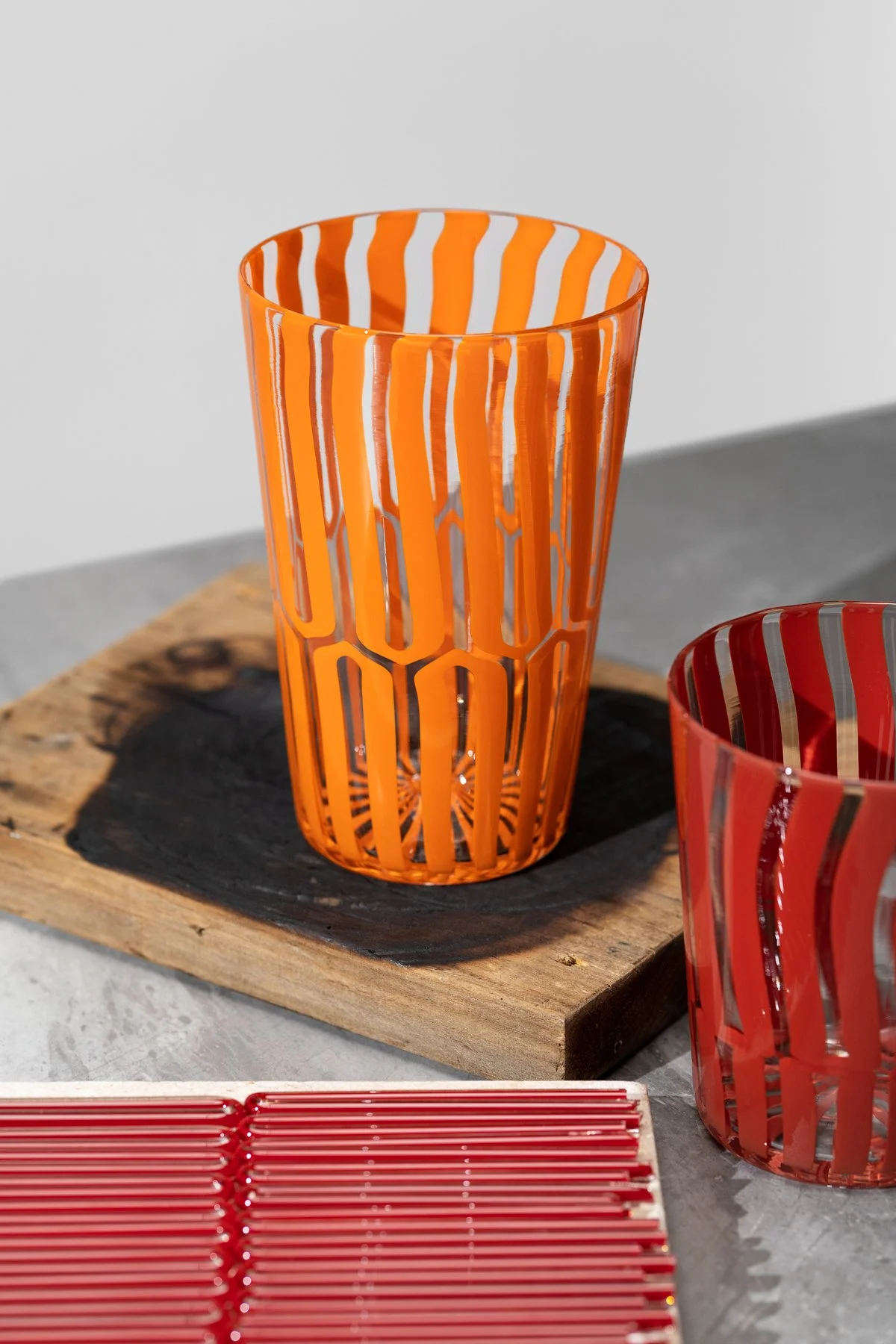 An orange striped glass Xander D'Ambrosio cup on a charred piece of wood next to a red striped cup. Glass cane in the foreground. Image by Loam