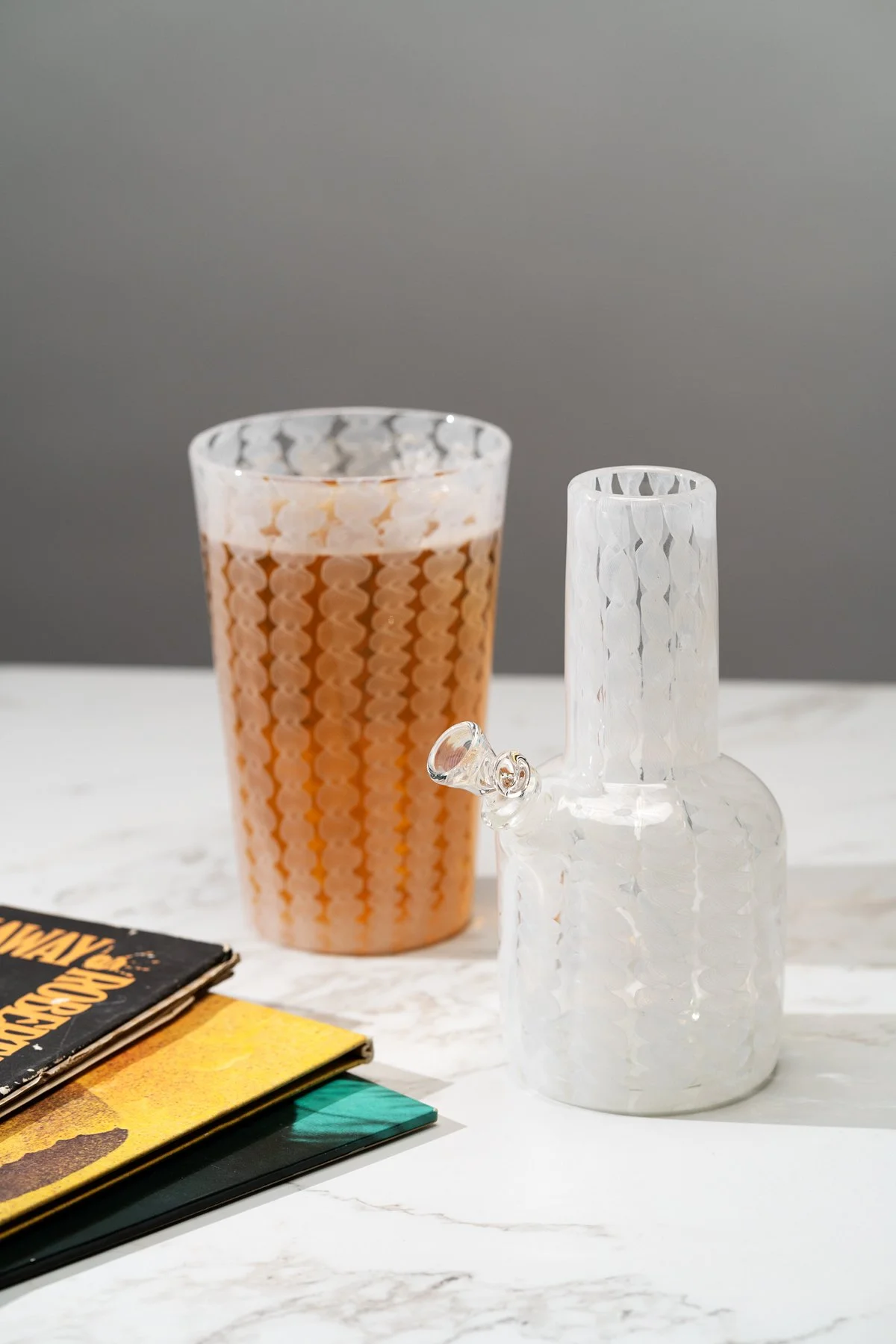 A clear and white glass Xander D'Ambrosio cup and water pipe set against a grey background with records peeking in. Image by Loam