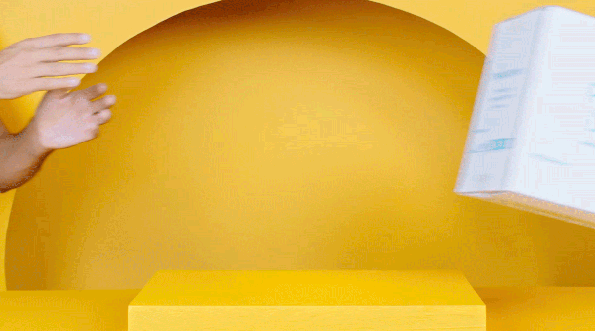 A yellow background with a yellow table, with a person's hand reaching towards a white box that is floating in mid-air. Sureal set for a PrettyLitter advertising video. 