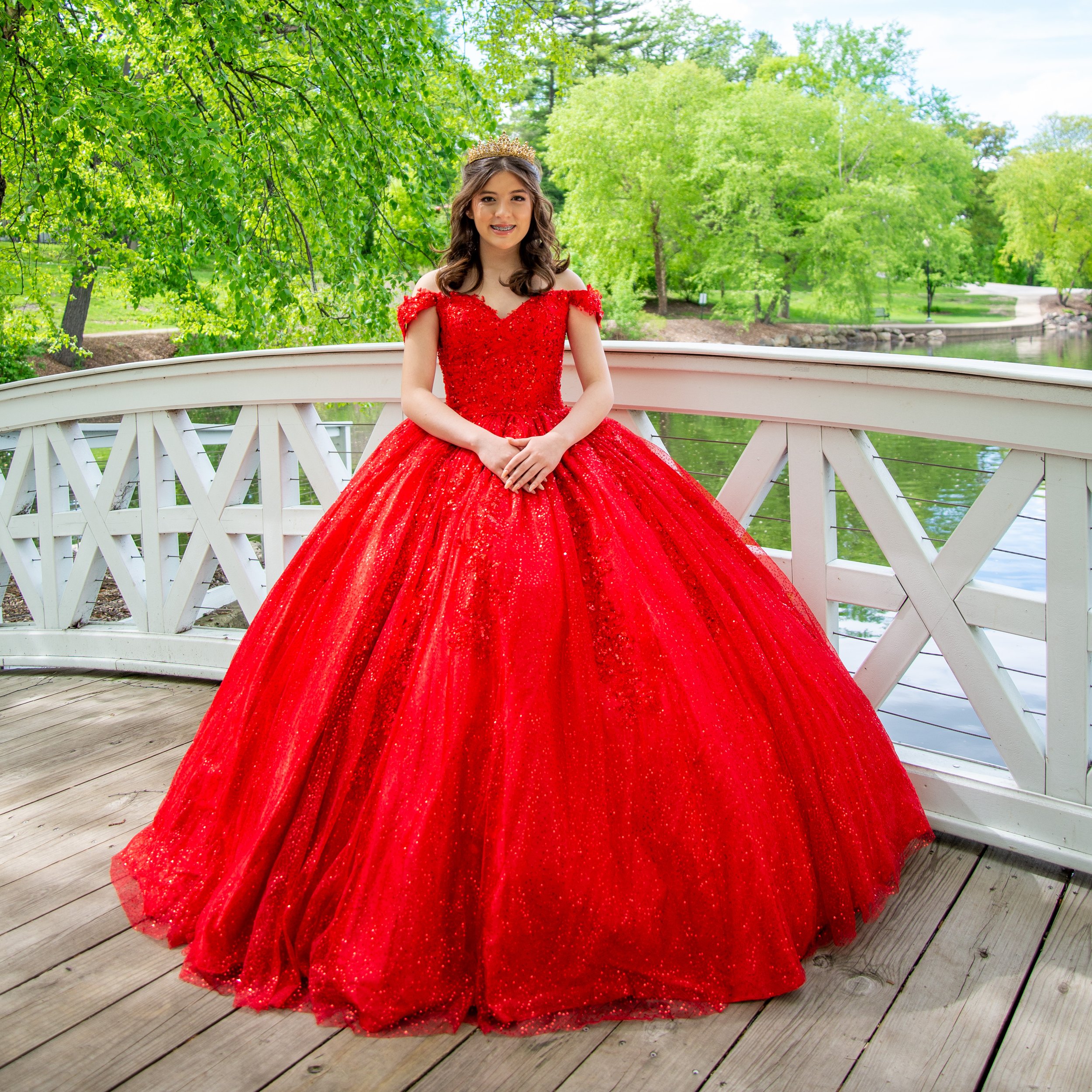 Young girl in a red ball gown standing on a white wooden bridge in a park.
