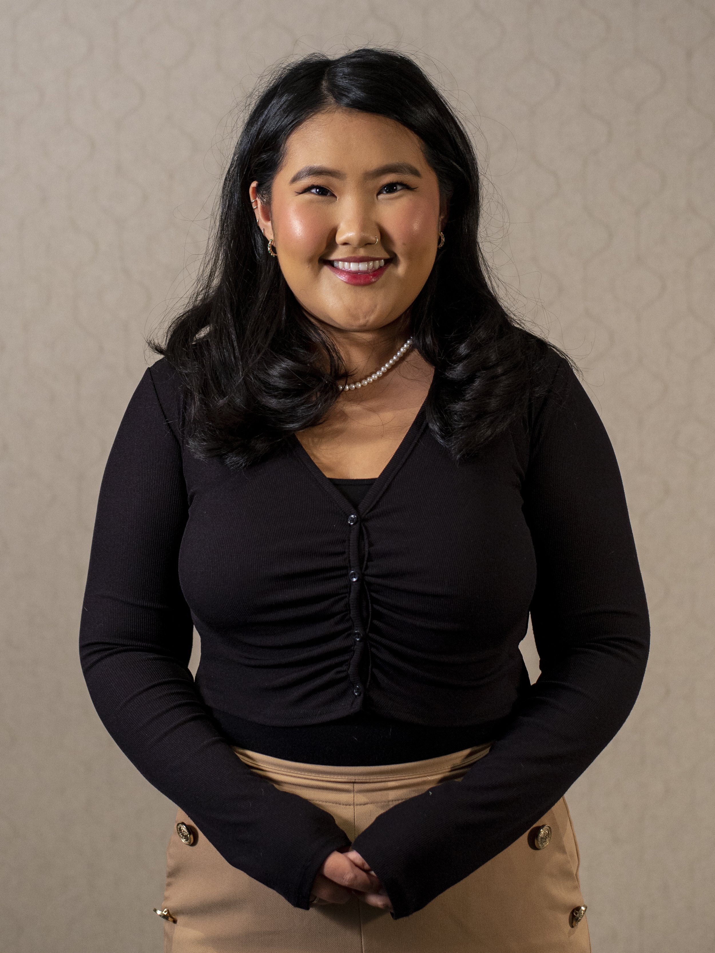 Smiling person wearing a black top and beige pants with button details, against a patterned background.