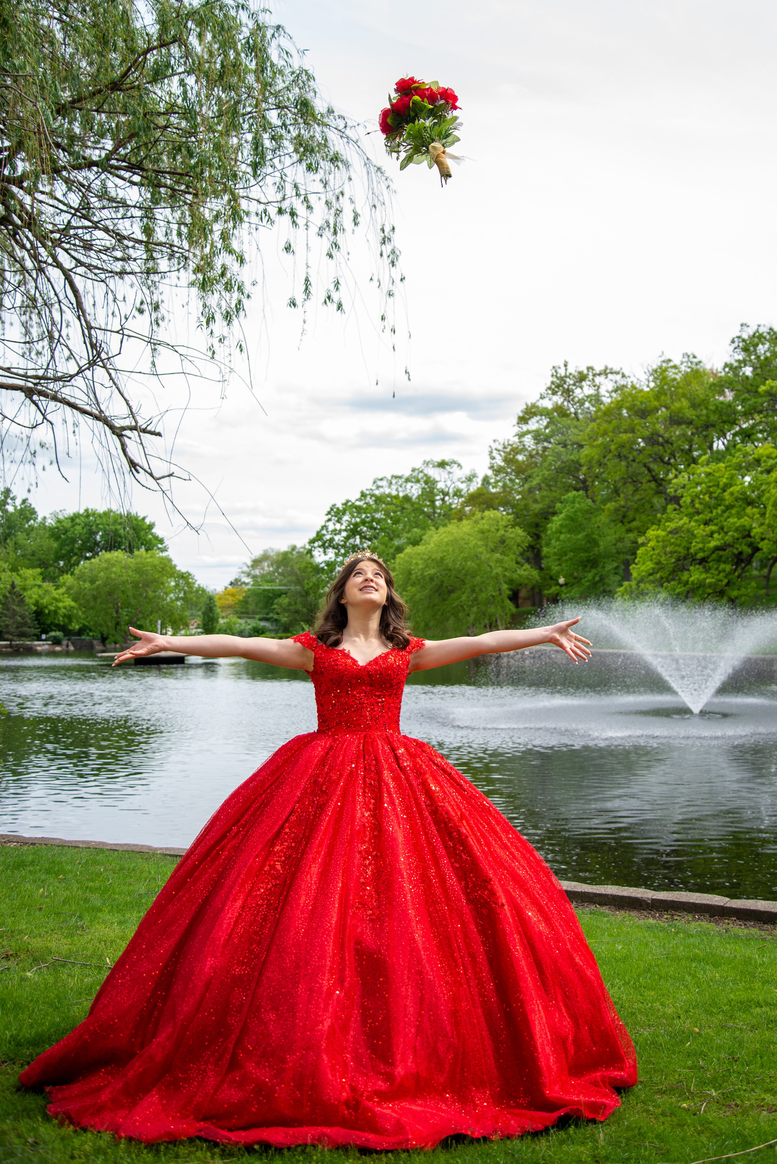 Young woman in a sparkling red dress tossing a bouquet into the air near a lake with a fountain and trees in the background.