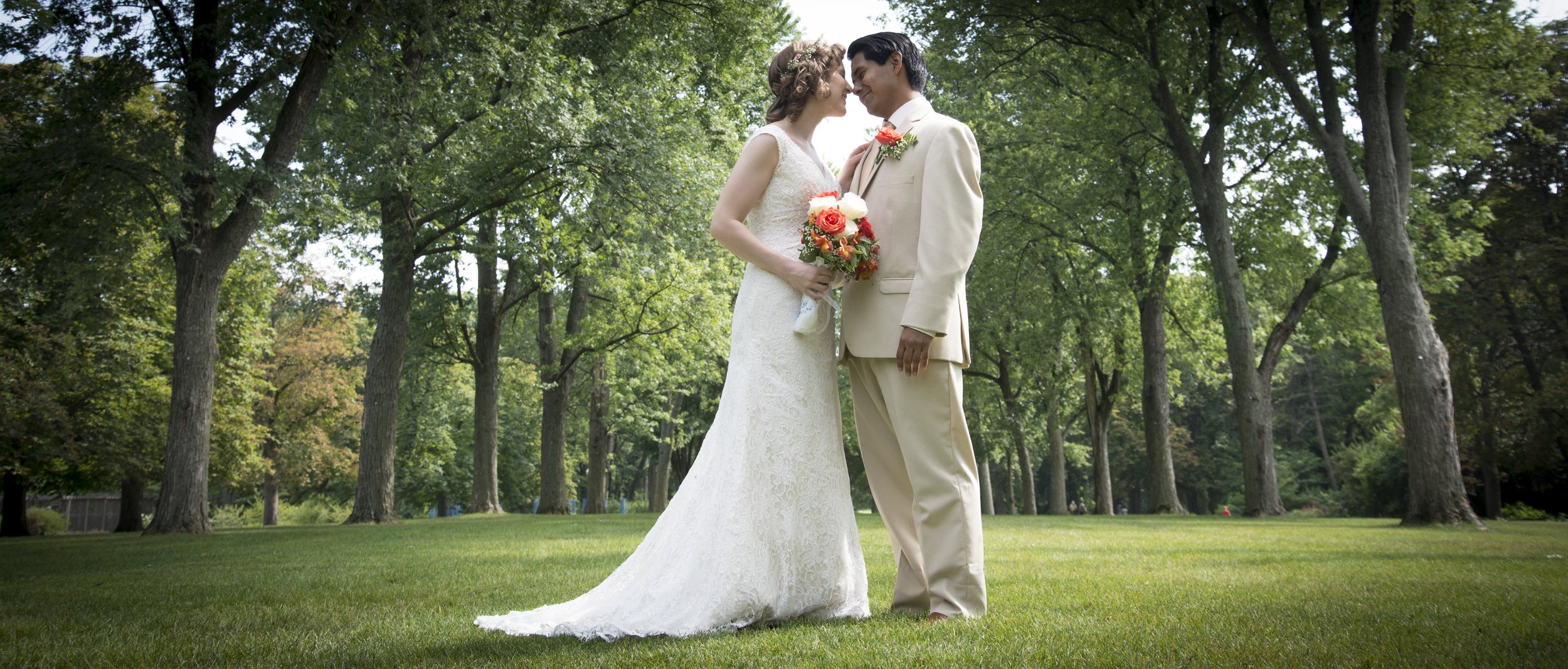 Bride and groom in a forest, bride in white dress holding bouquet, standing on grass.