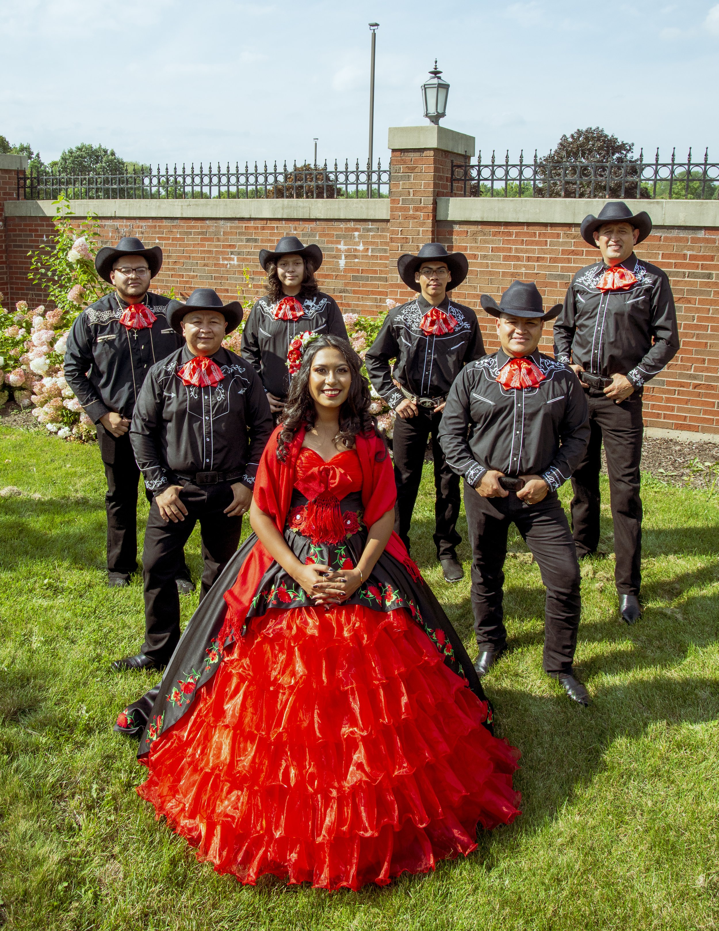 A woman in a bright red traditional dress stands in front of six men wearing black outfits with red bow ties and cowboy hats, posing outdoors on a green lawn with a brick wall and flowers in the background.