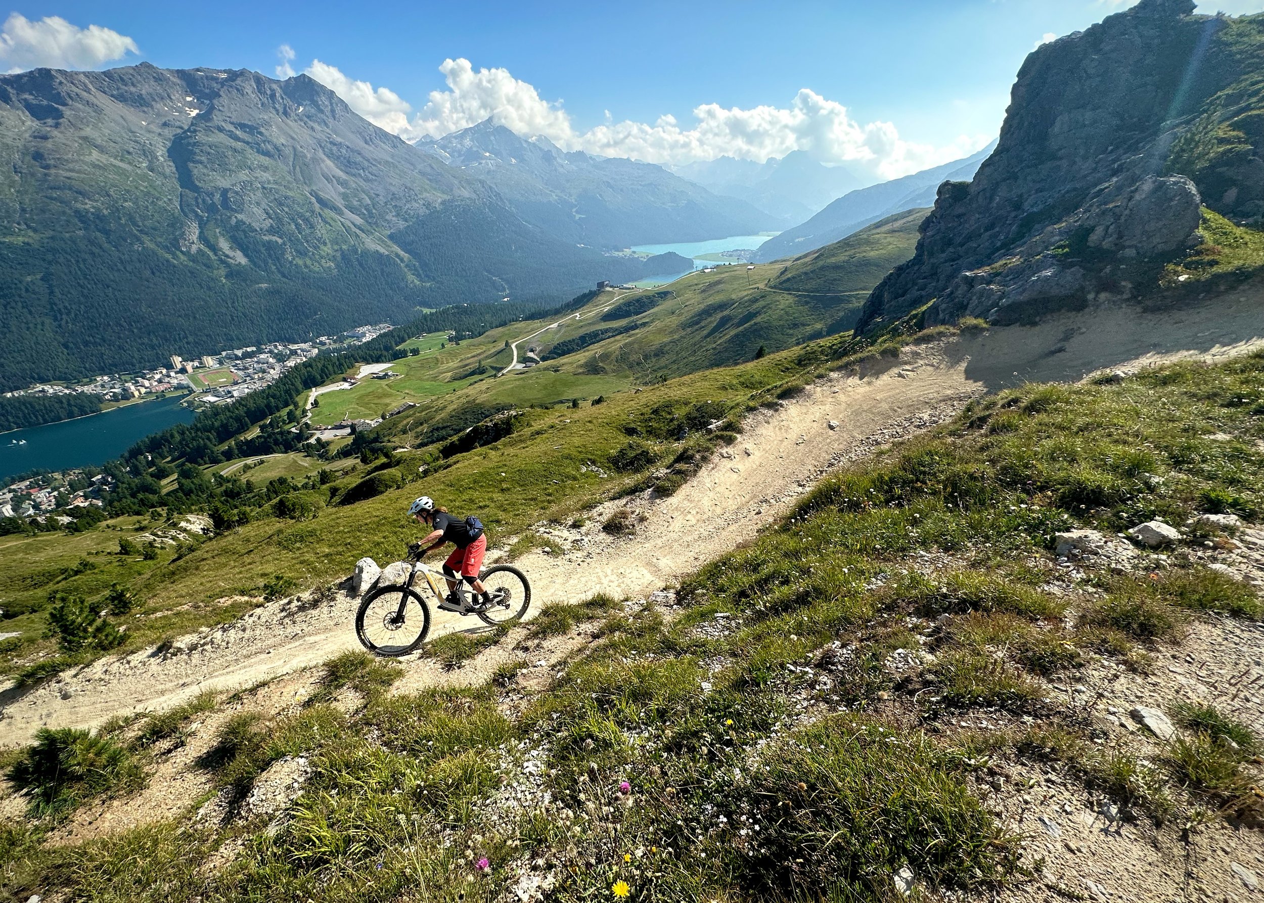 A person mountain biking downhill on a dirt trail in a mountainous landscape with lakes and a town below.
