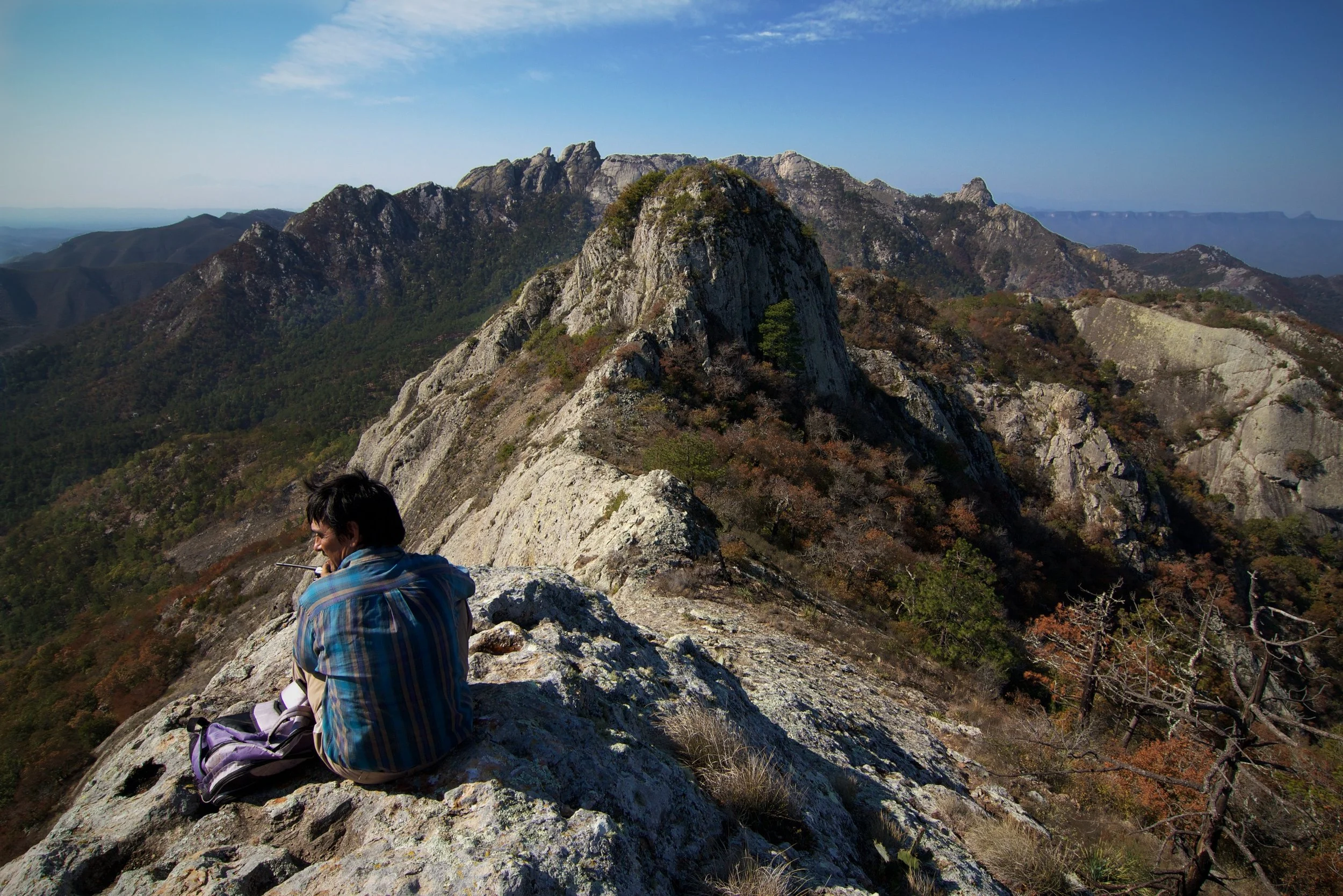 A person is sitting on a rocky mountain ledge with a mountain range and blue sky in the background.