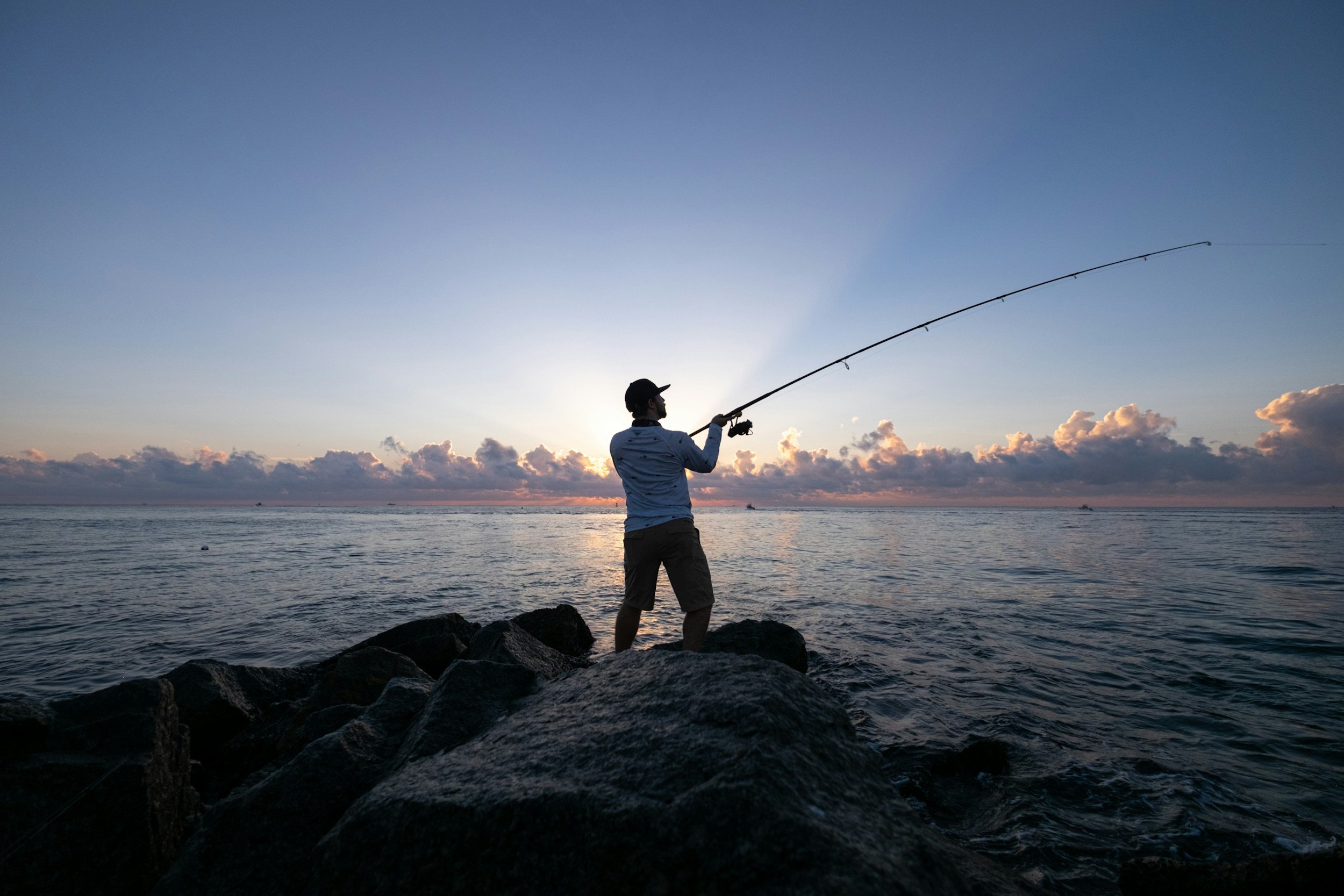 A man standing on rocks fishing outdoors as the sun sets