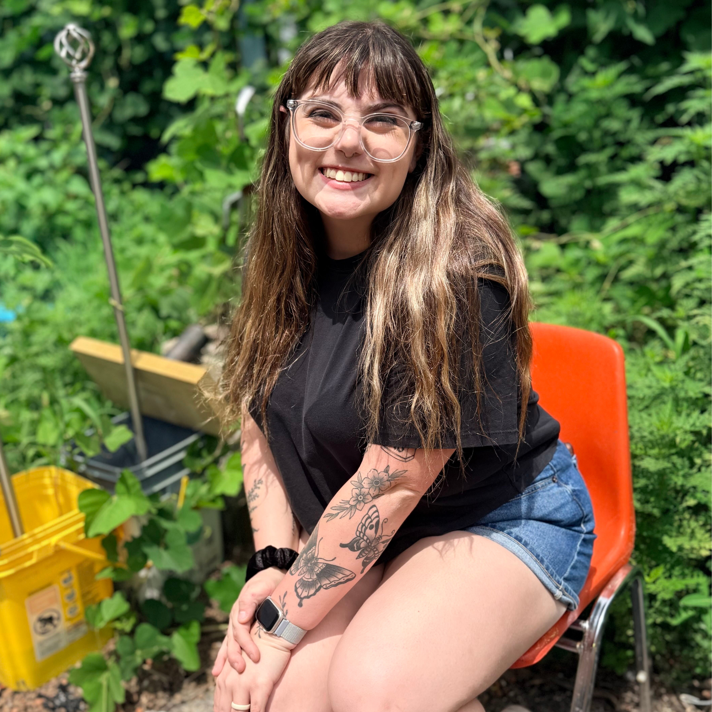 A woman with tattoos, glasses, and long brown hair smiling outdoors while sitting on a colorful chair in a garden or farm setting, with green plants and yellow containers around.