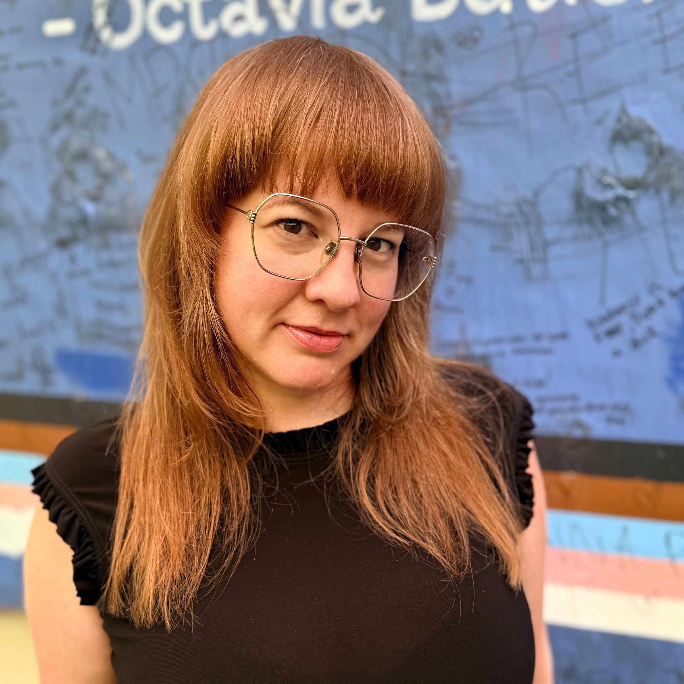 Close-up of a woman with red hair and glasses, wearing a black shirt, smiling, with a blue wall with writing behind her.