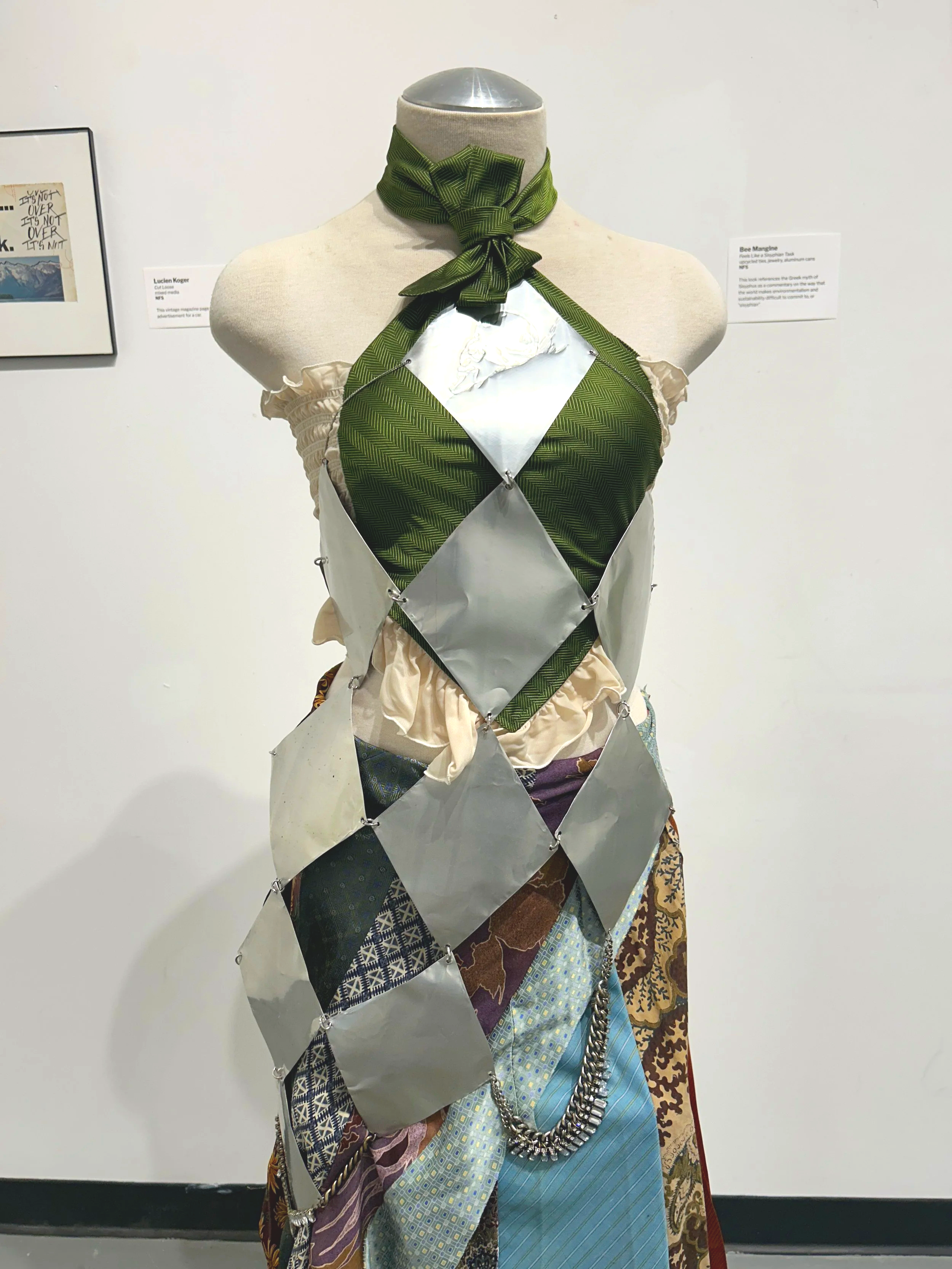 A dress made of various fabric swatches and mirrored diamond-shaped pieces, with a green bow tie around the neck, displayed on a mannequin against a white wall with framed artwork.