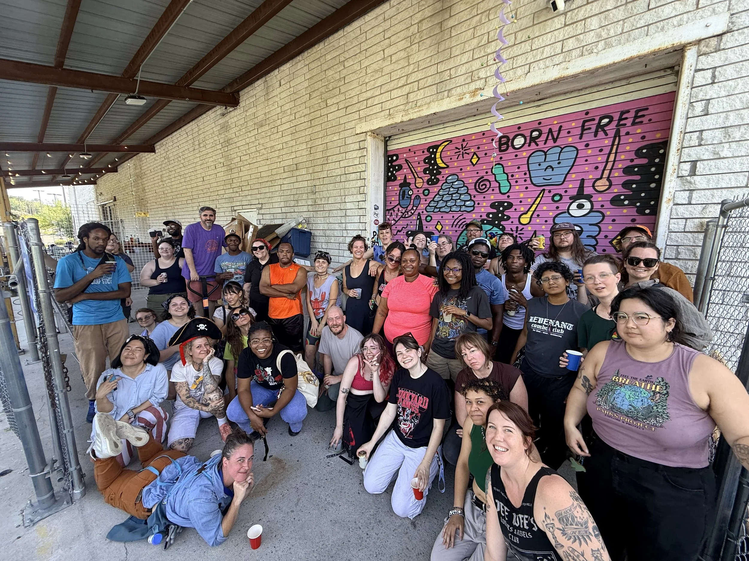 A large group of diverse people posing together for a photo outside under a covered area with a brick wall, colorful graffiti art, and string lights in the background.