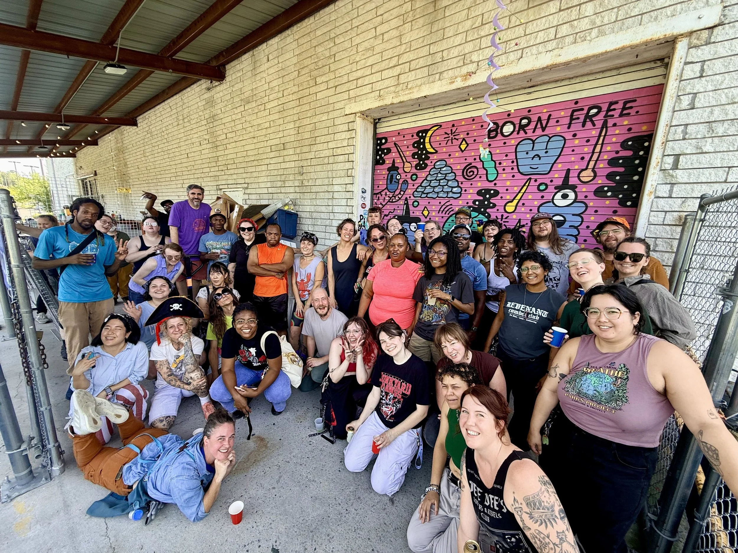 A group of diverse people gathered under a covered area near a wall with graffiti art that says "BORN FREE". They are smiling, holding drinks, and some are making peace signs.