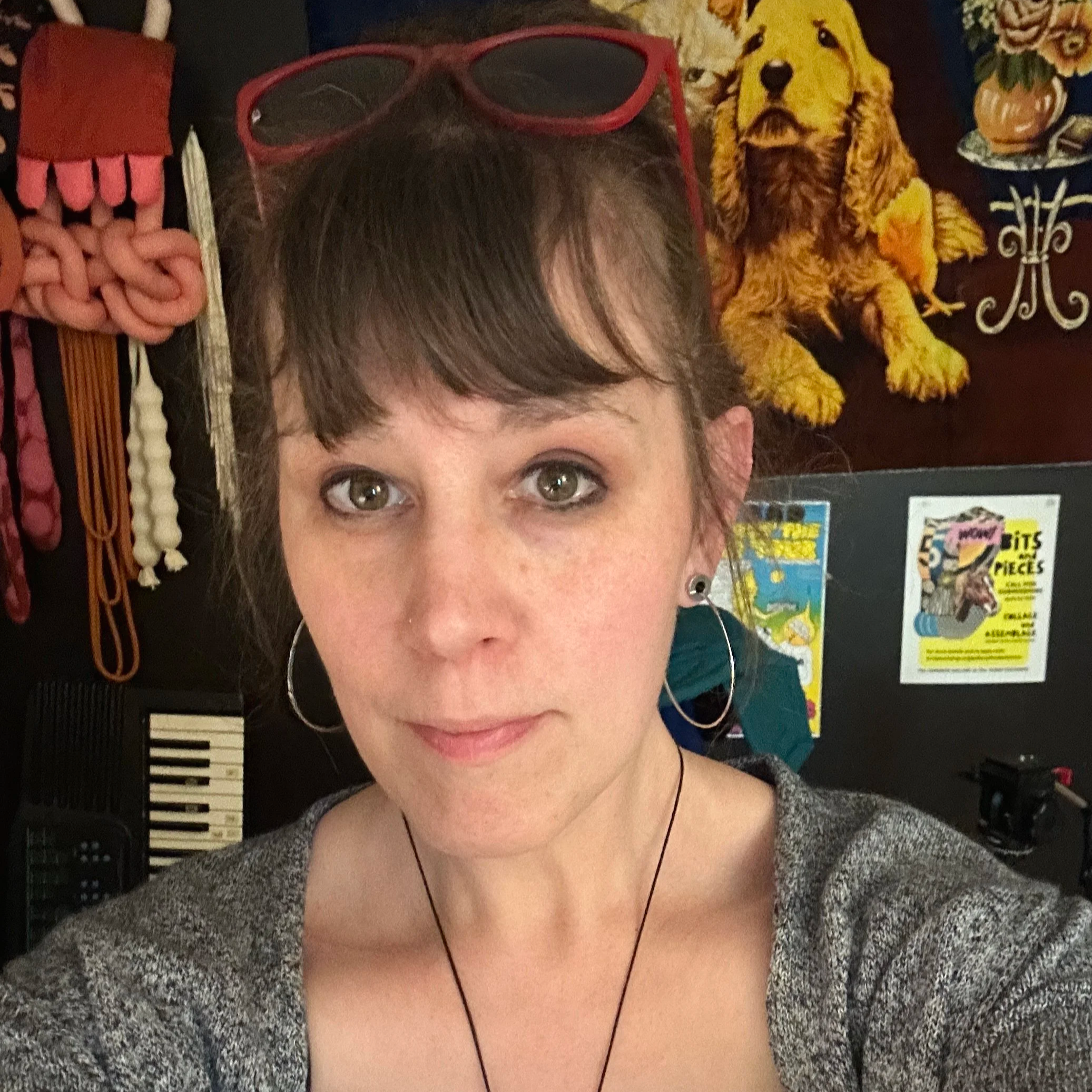 Close-up of a woman's face with brown eyes and short dark hair, wearing red glasses on her forehead, with a backdrop of books and colorful posters.