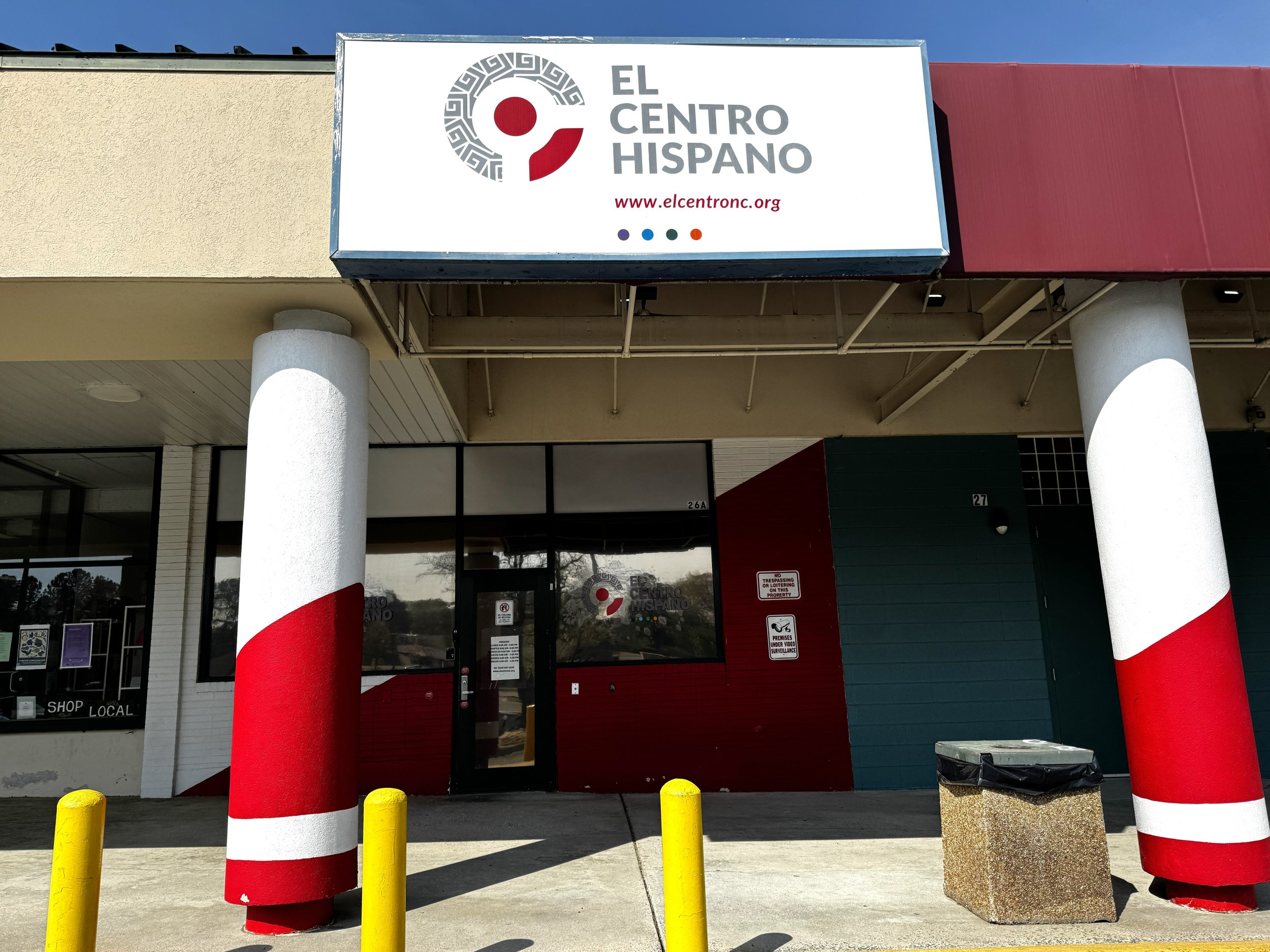Sign for El Centro Hispano above the entrance of a building, with red and yellow protective bollards in front.