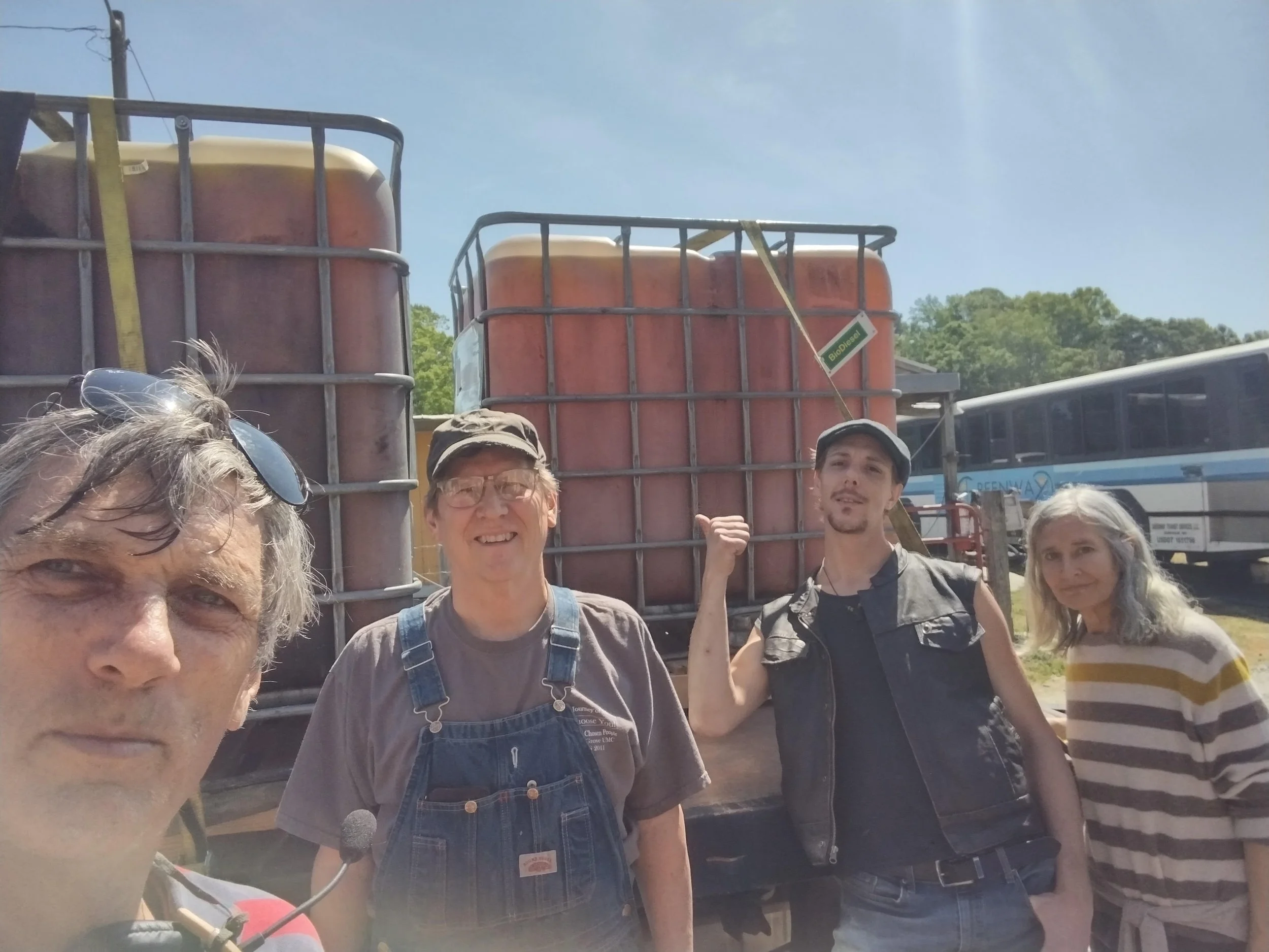 A group of people outdoors, standing in front of large orange containers labeled 'BioDiesel.' They are smiling and making fist gestures, with trees, a bus, and a light blue sky in the background.